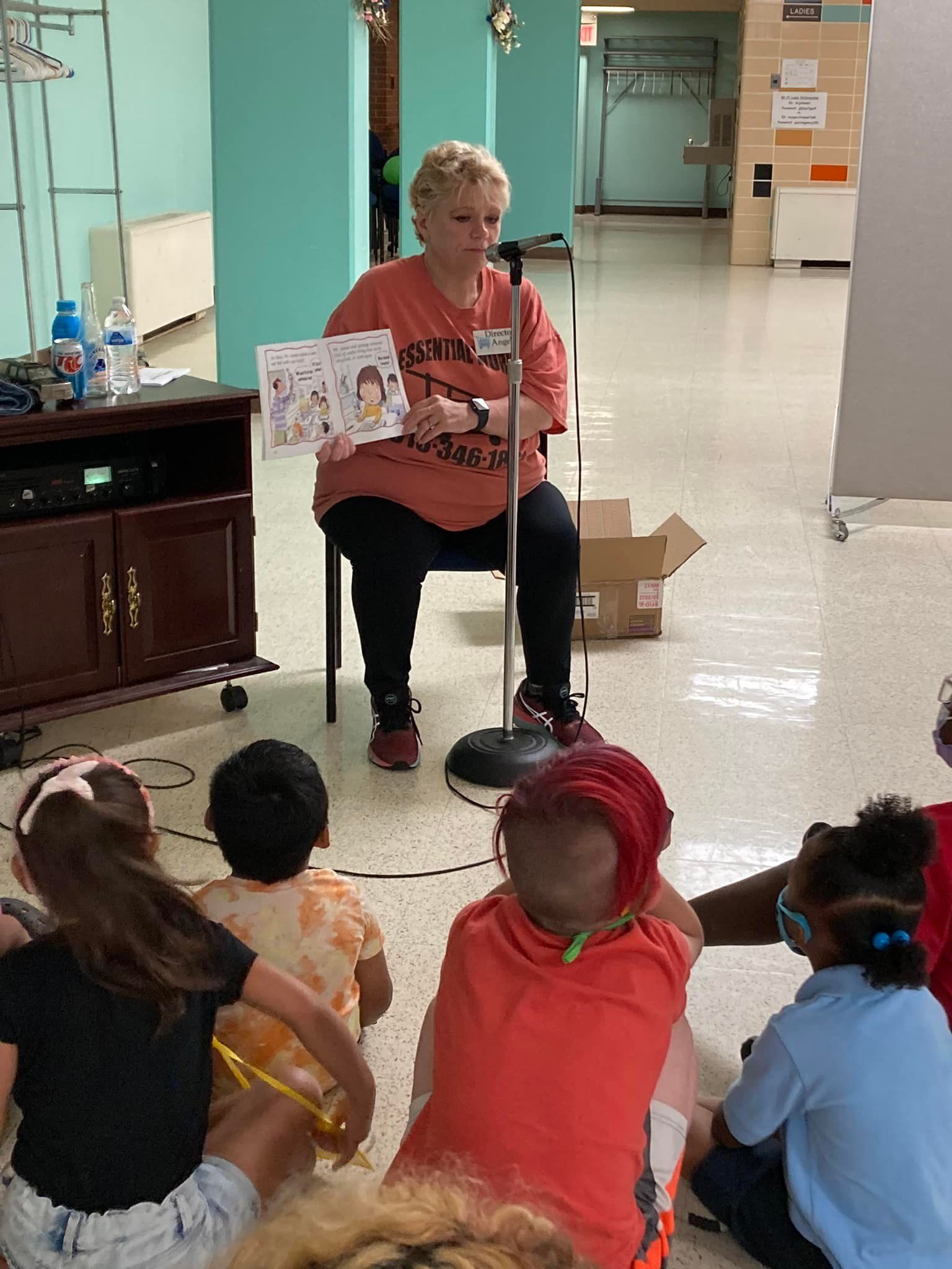 Teacher reading to seated children in a hallway, holding an illustrated book and puppet.