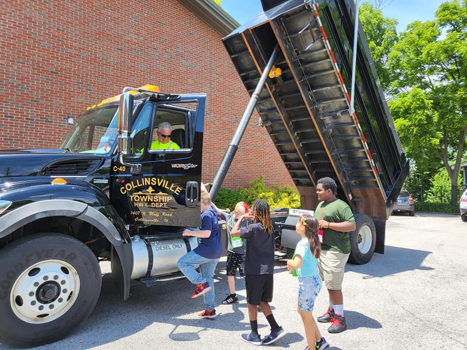 Children gather beside a black dump truck with its bed raised near a brick building on a sunny day