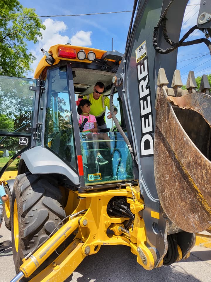 Two workers in a yellow DEERE backhoe at a construction site under a blue sky