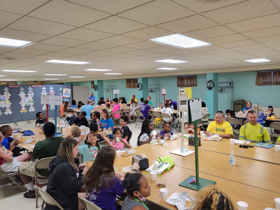 Crowded cafeteria with many people seated at long tables, eating and talking under bright ceiling lights.