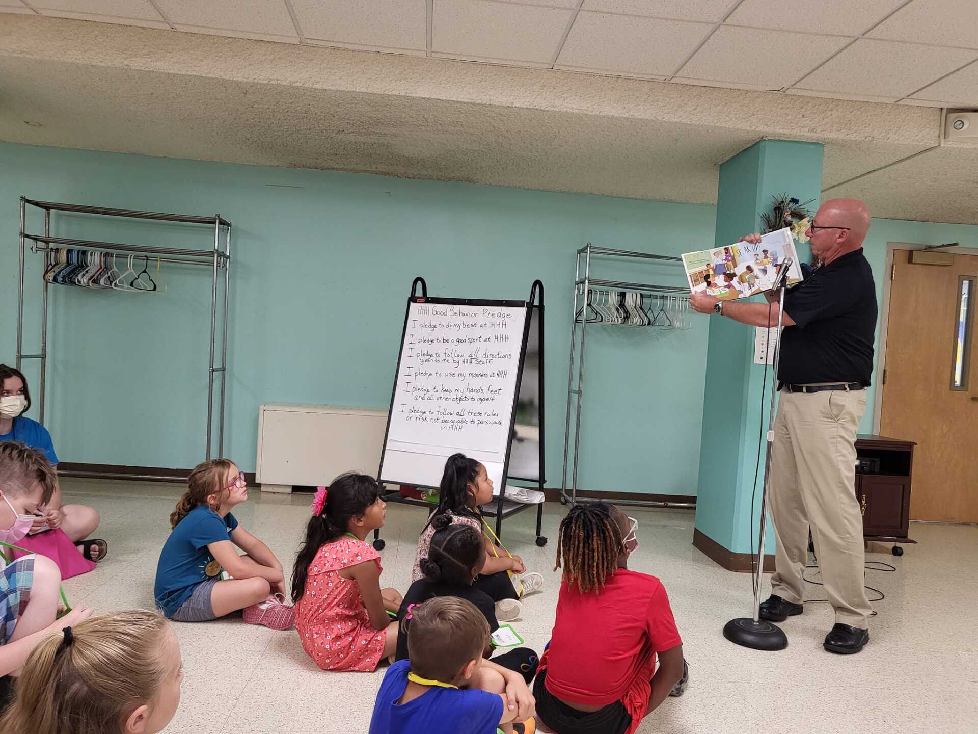 Teacher reads a book to seated children in a classroom with a whiteboard and coats on racks.