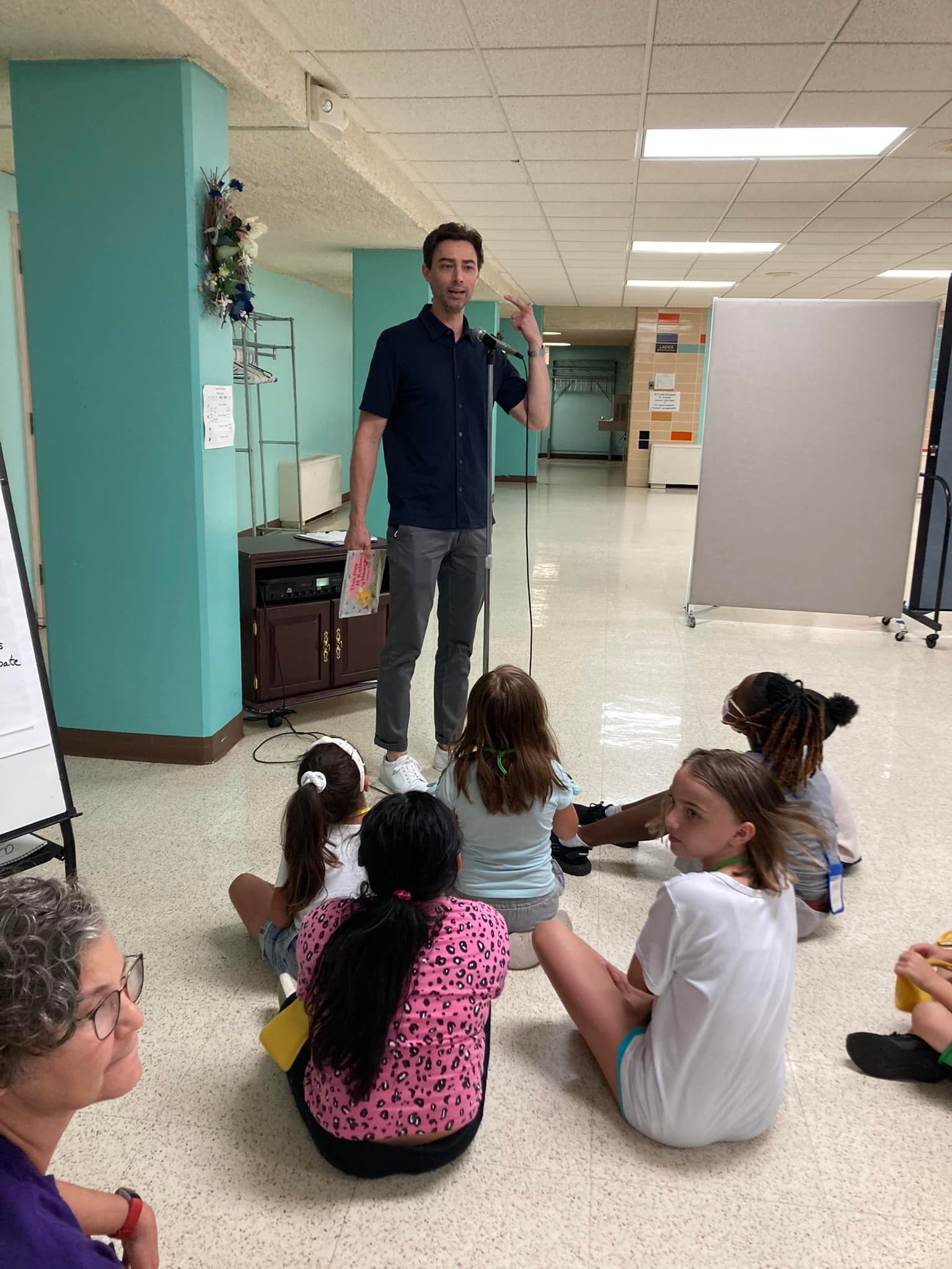 Man speaking to seated children in a school hallway during a class discussion