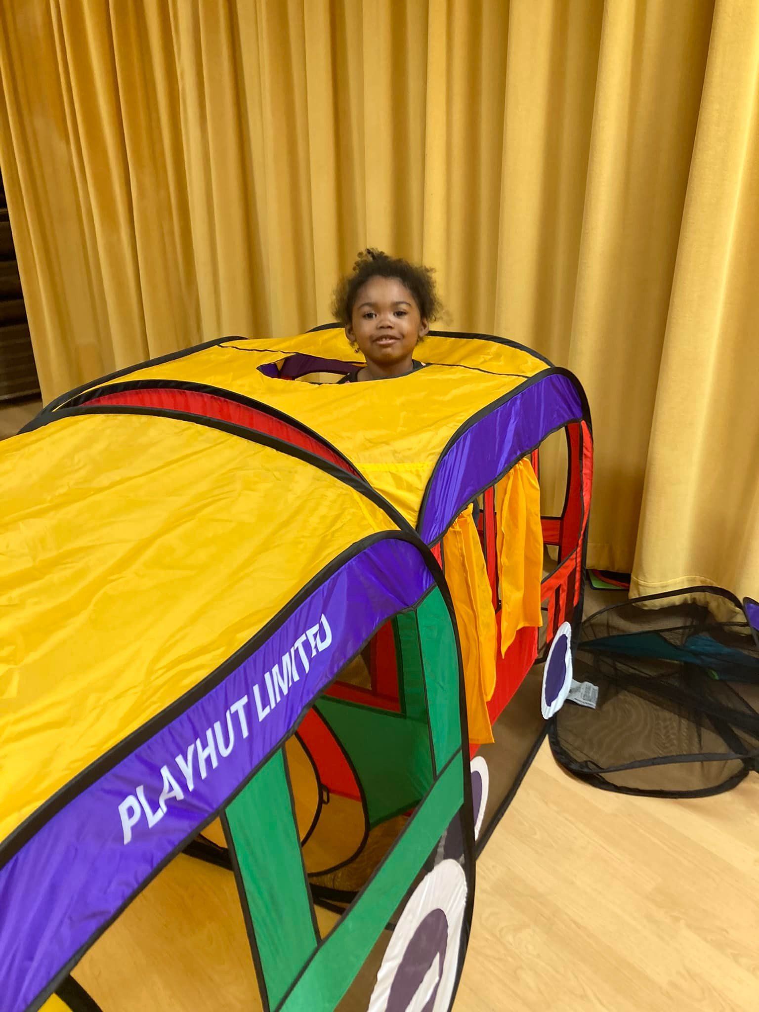 Child peeking out of a colorful play tunnel in a room with yellow curtains