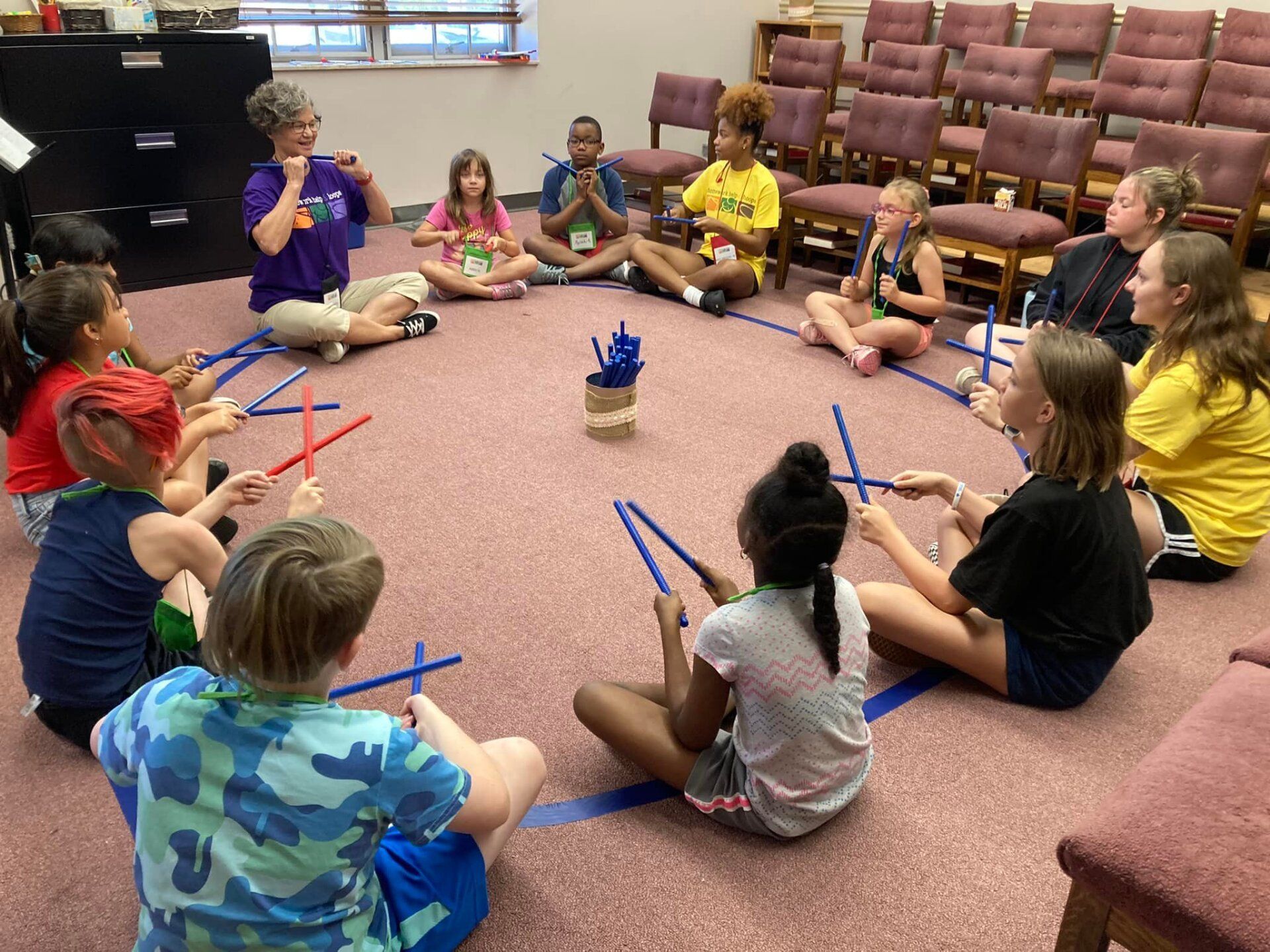 Children seated in a circle on a carpet, holding colorful rhythm sticks in a classroom activity