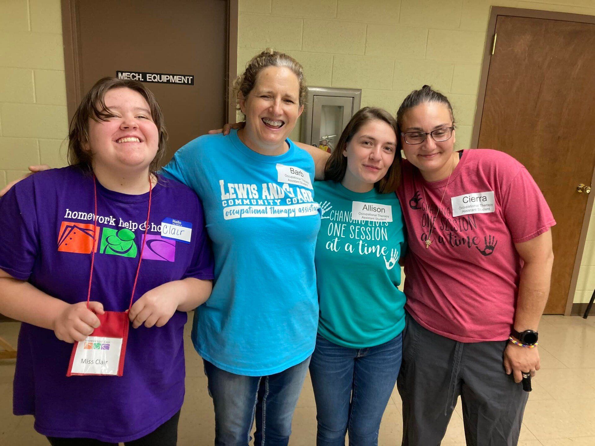 Four people smiling indoors, standing shoulder to shoulder in colorful T-shirts and name tags.