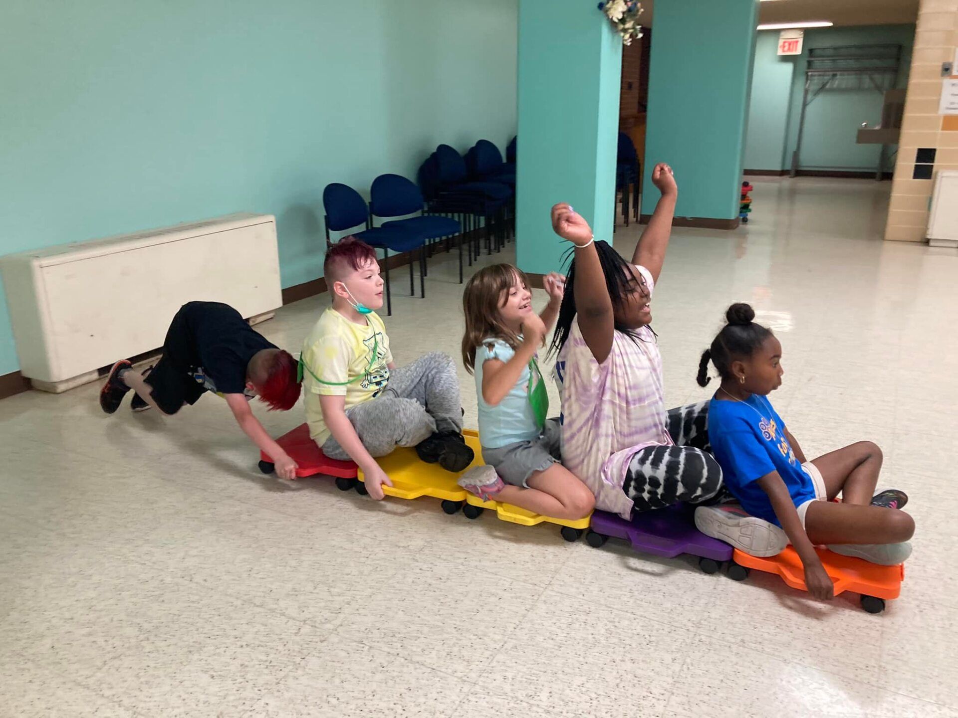 Children sitting in a row on colorful mats in a classroom, one raising a hand.