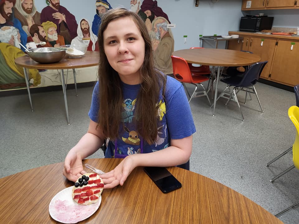 Person seated at a table, holding a plate with food in a classroom setting.