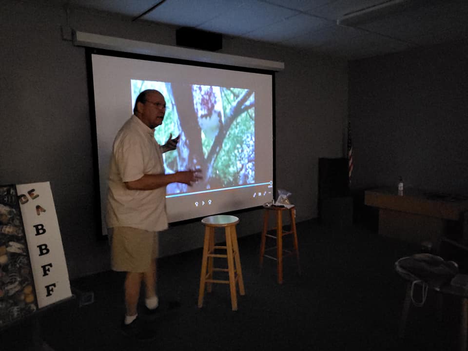 Man presenting beside a projected slide in a dim room, with stools and a vertical ABBF sign nearby