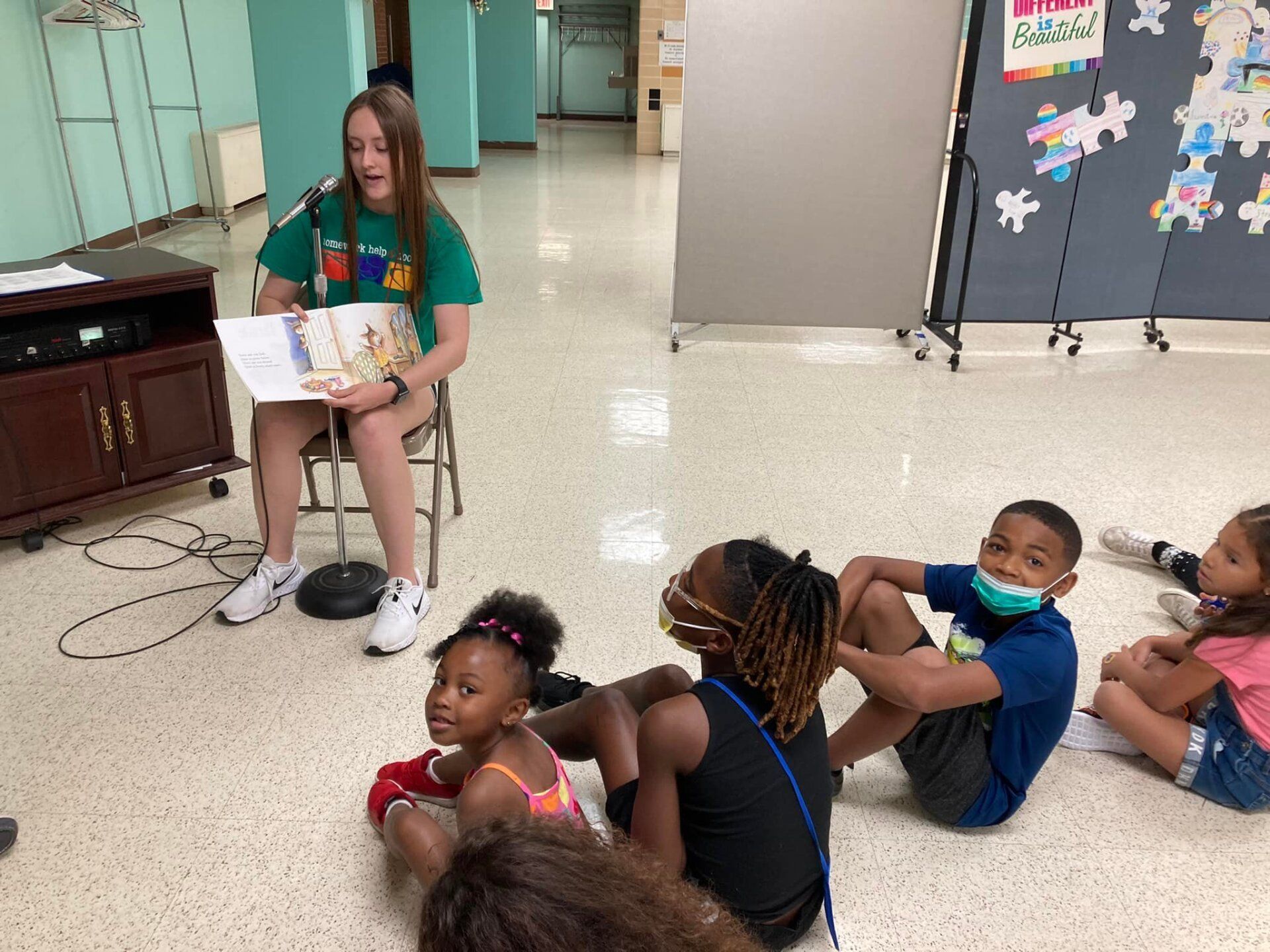 Children sitting on a school hallway floor near a girl seated on a stool holding a book