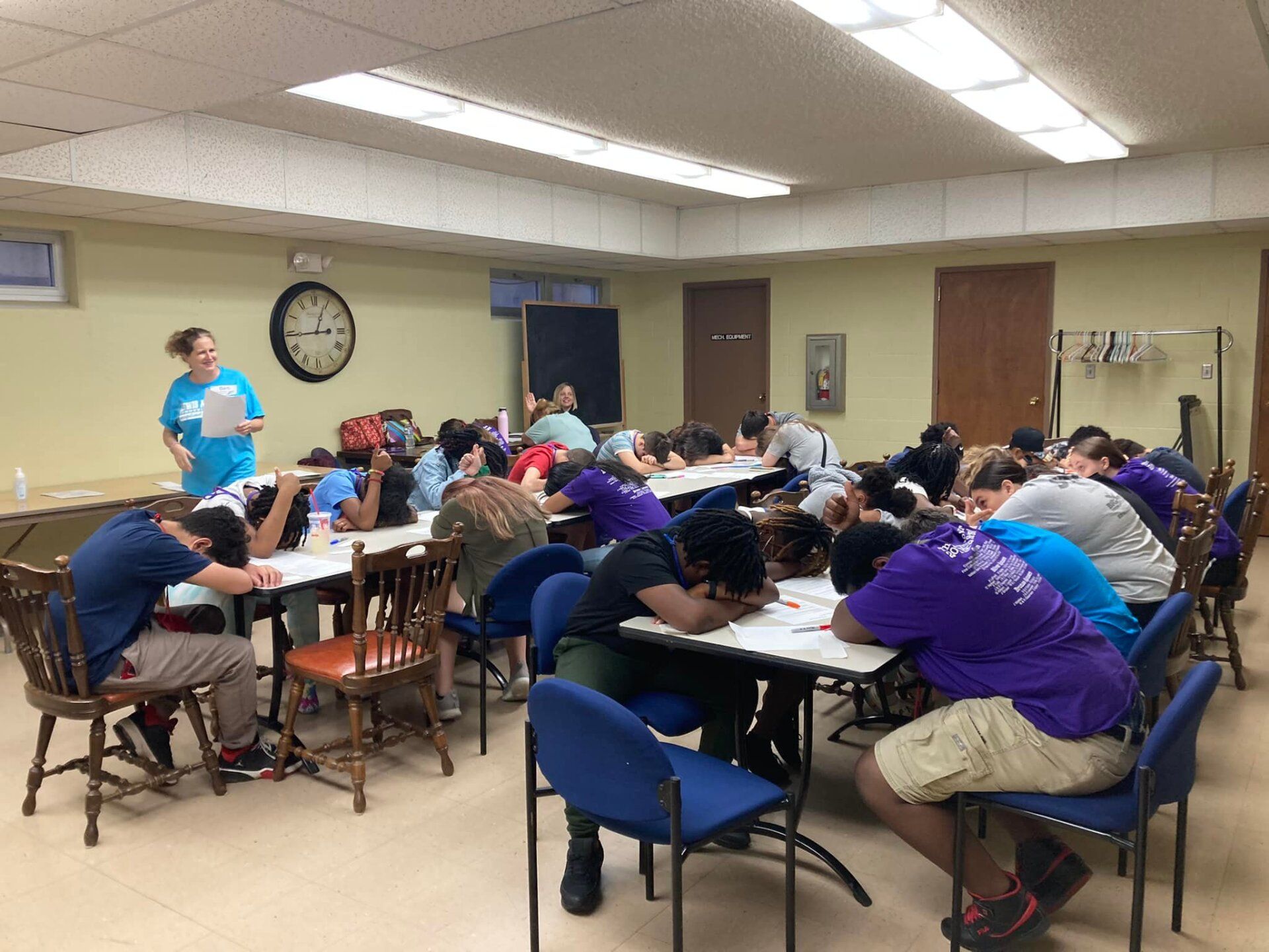 People seated at tables in a classroom, leaning forward with heads down during an activity.
