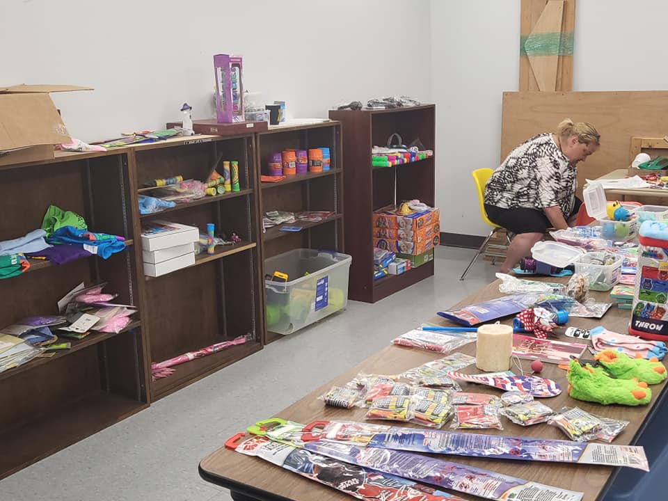 Woman kneels beside tables covered with craft supplies in a small room with shelves of toys and materials.