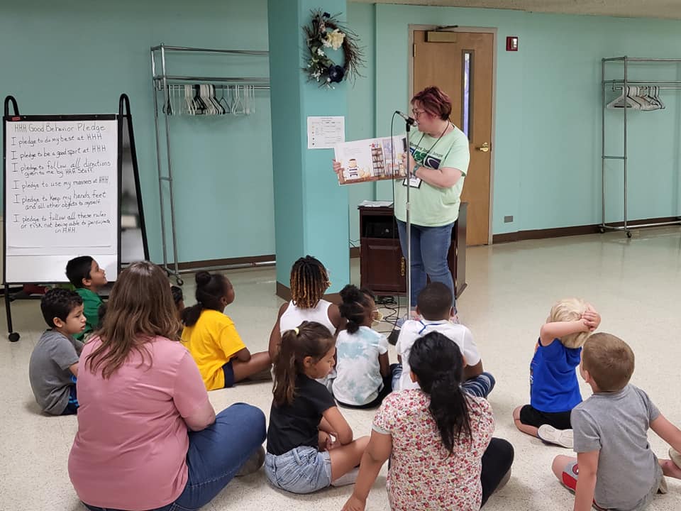 Teacher reads to seated children in a mint-green classroom with whiteboards and wall hooks.