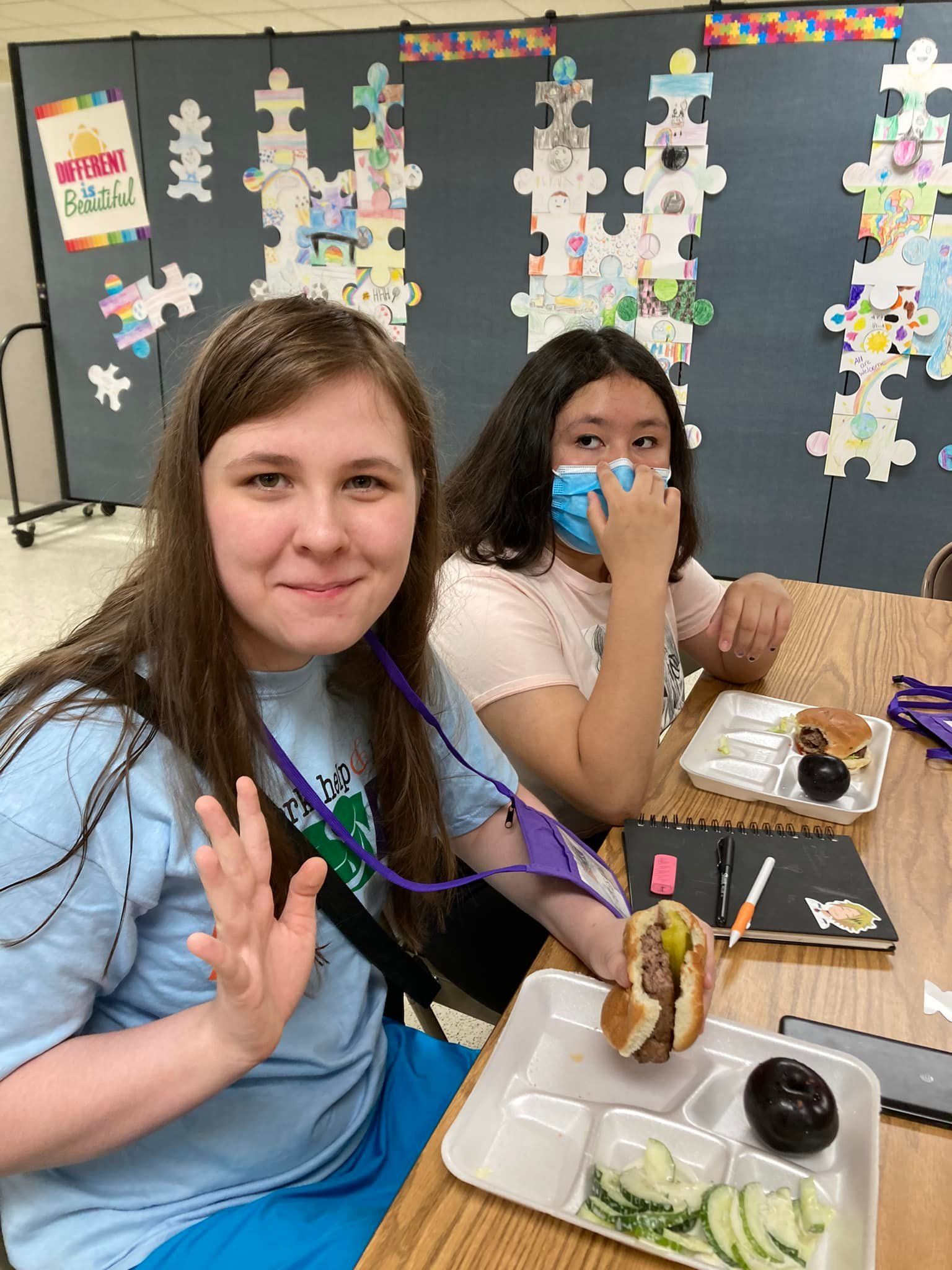 Two students at a cafeteria table, one waving and one wearing a blue mask, with lunch trays and decorations behind them