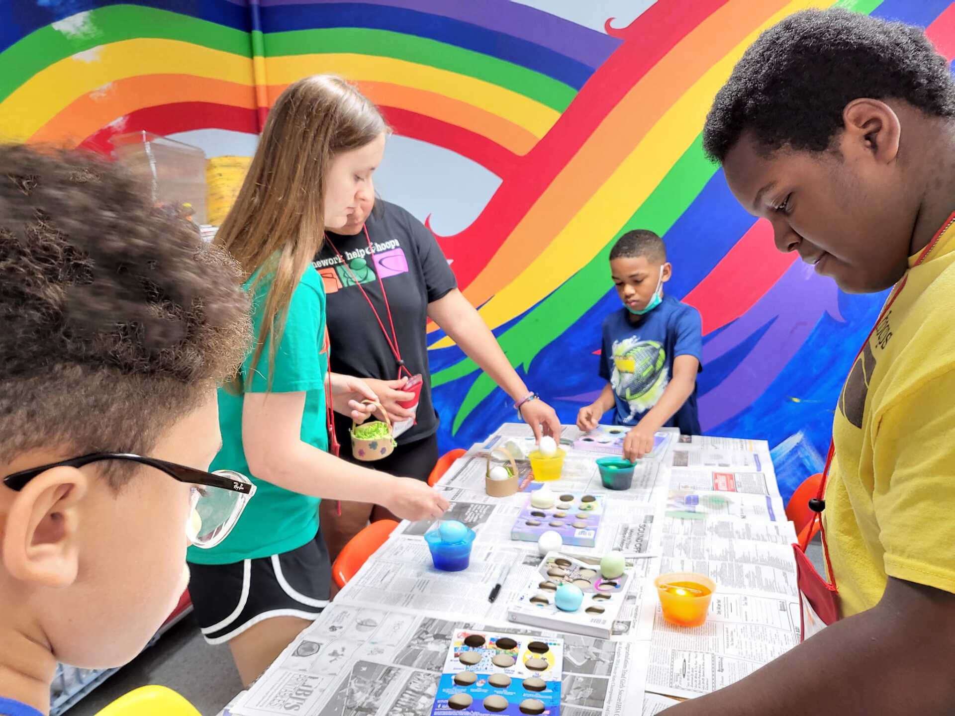 Children mixing art supplies at a table in front of a colorful rainbow mural