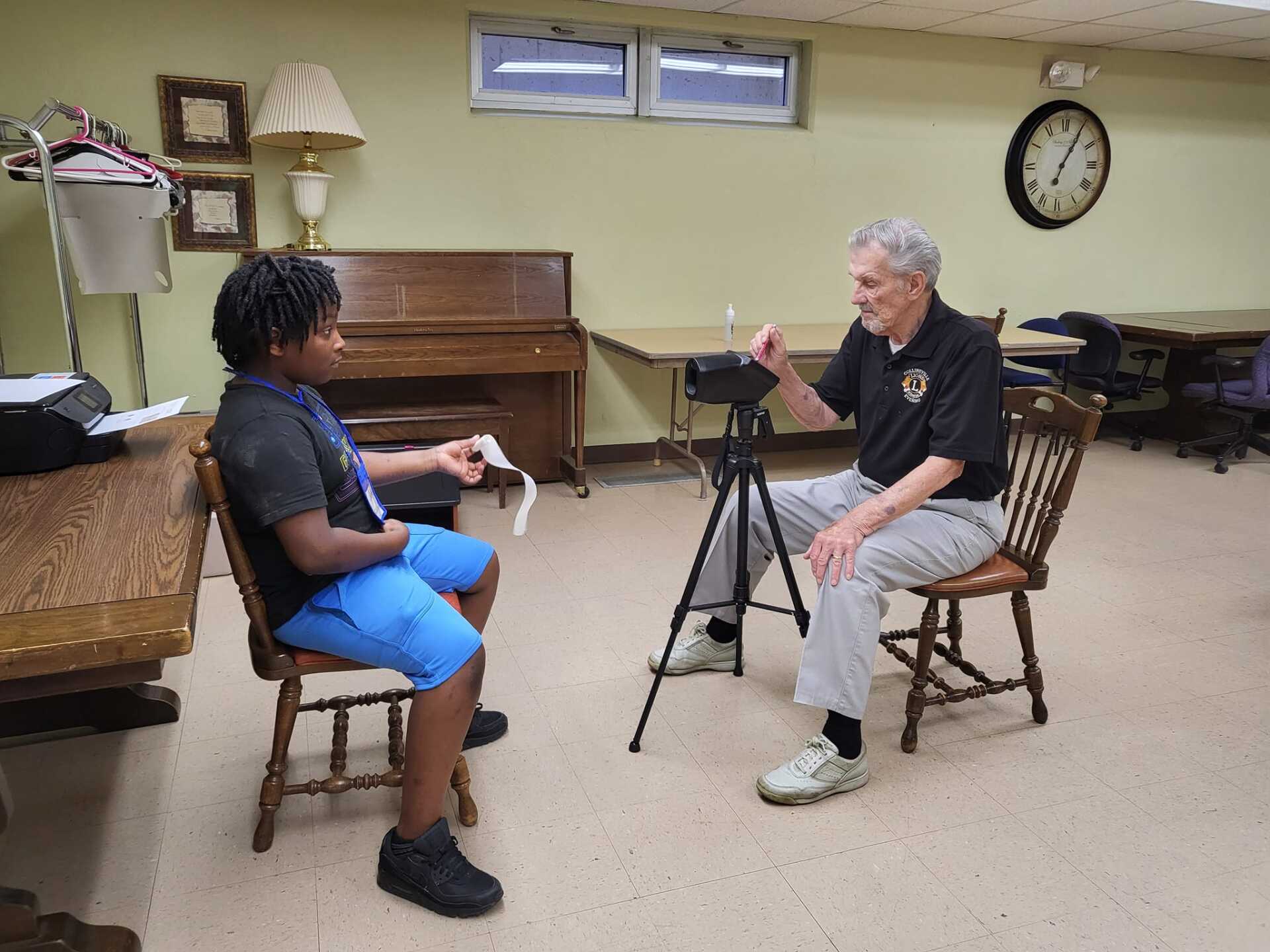 Two people seated in a room during an interview, with a tripod-mounted camera between them.