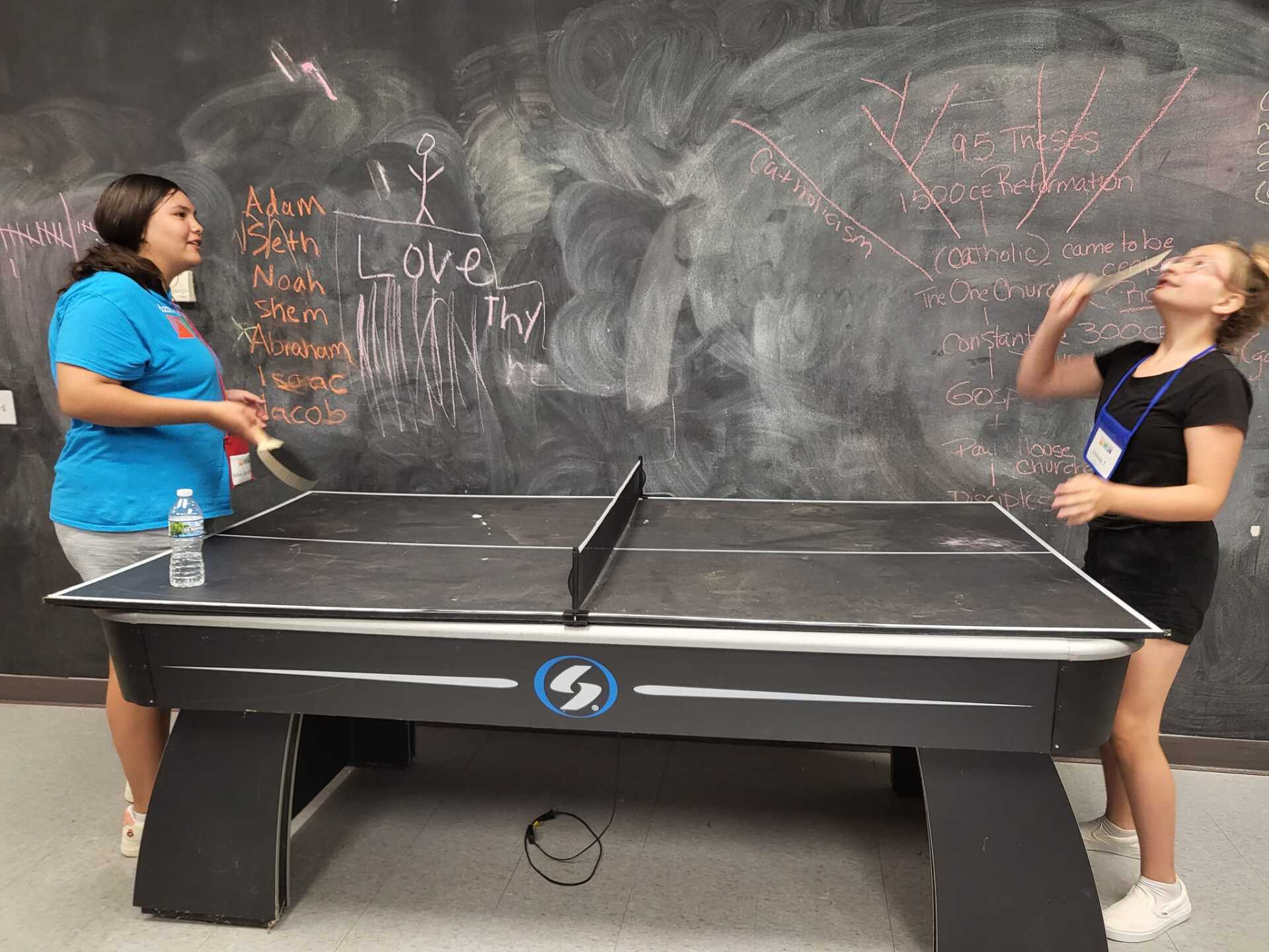 Two people playing ping-pong in front of a chalkboard wall covered with doodles and writing