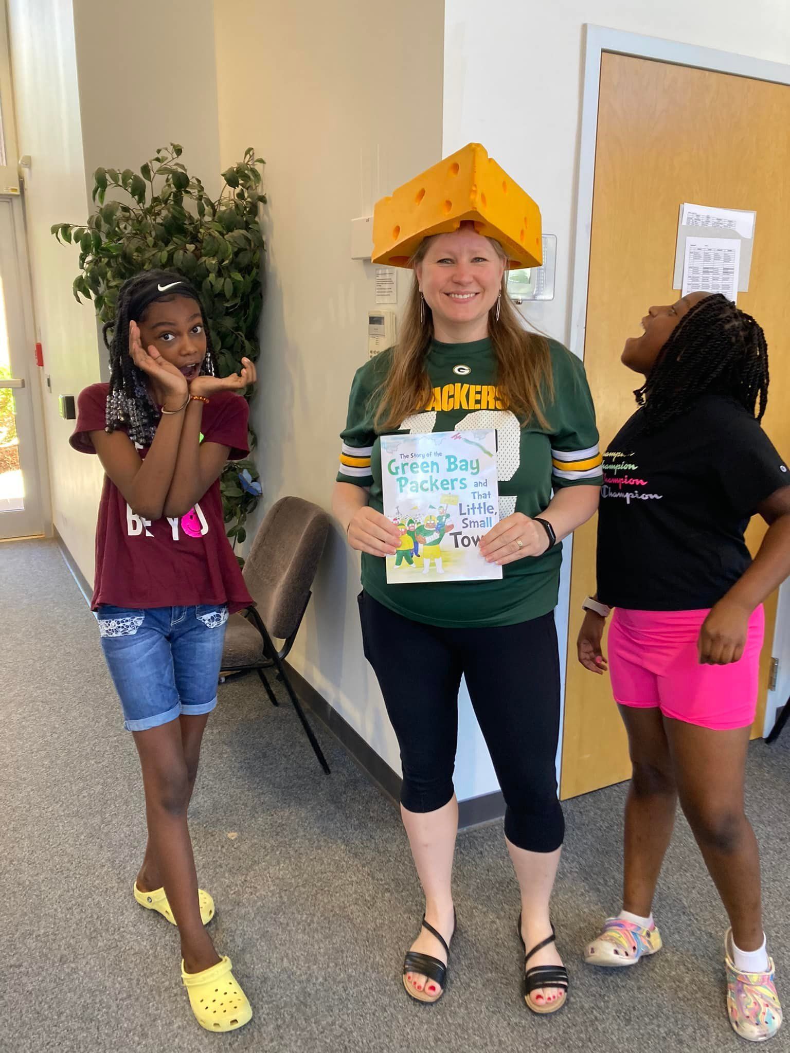Three children in a hallway; one holds a sign and wears a yellow paper hat.