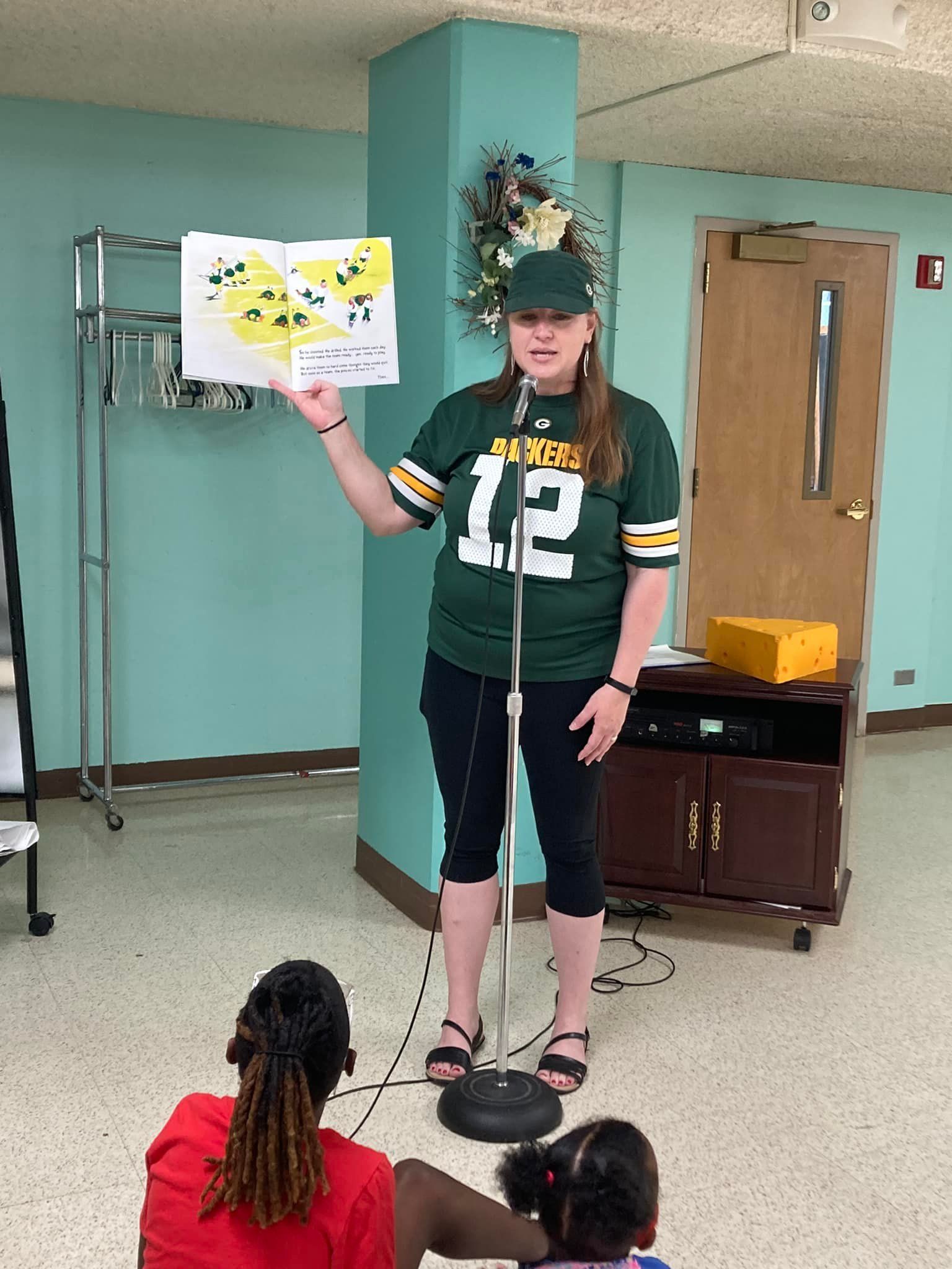 Person in a green jersey reads to children in a classroom, holding up a butterfly book.
