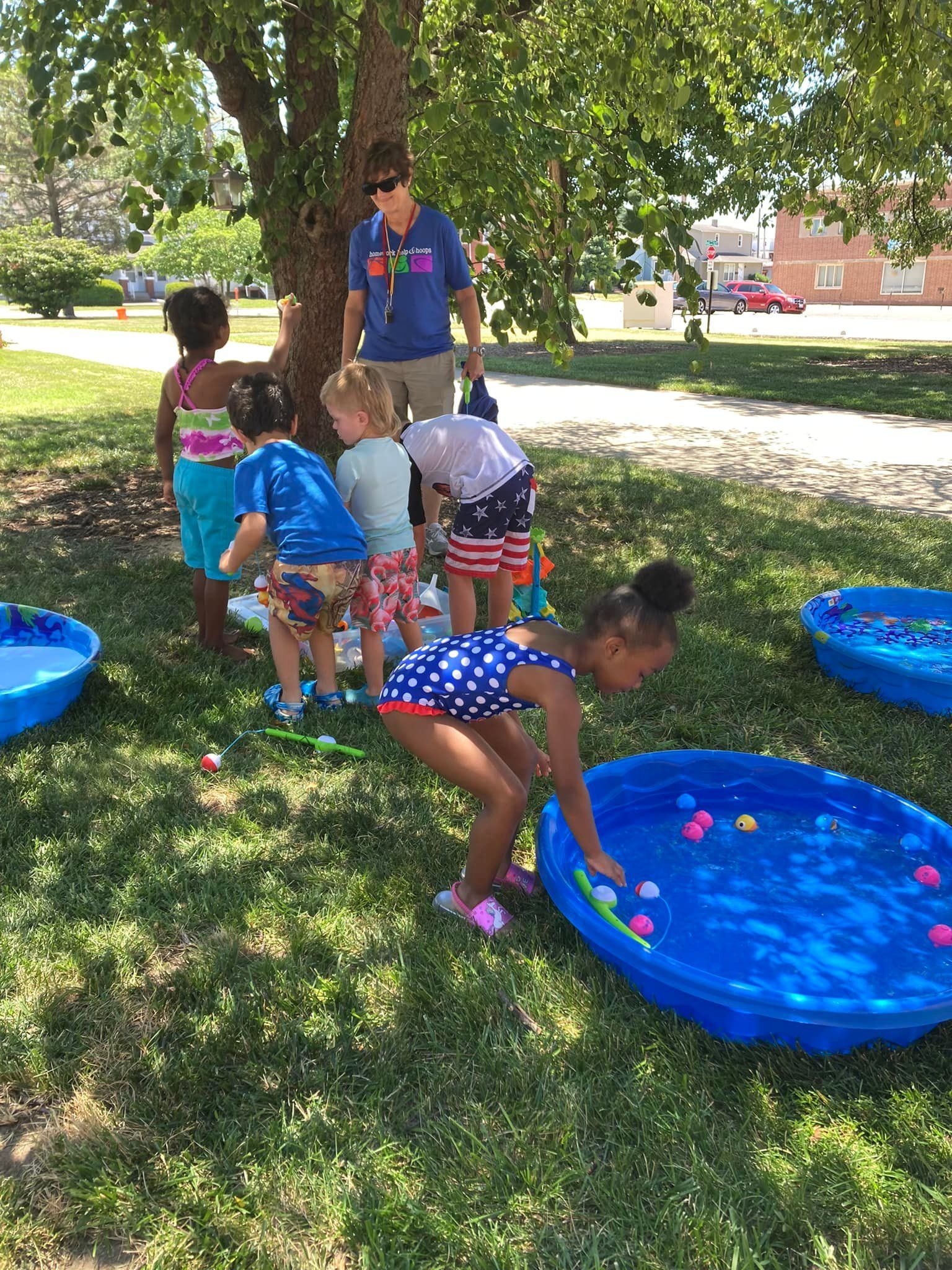 Children play with water toys in a grassy park near a person standing in the shade.