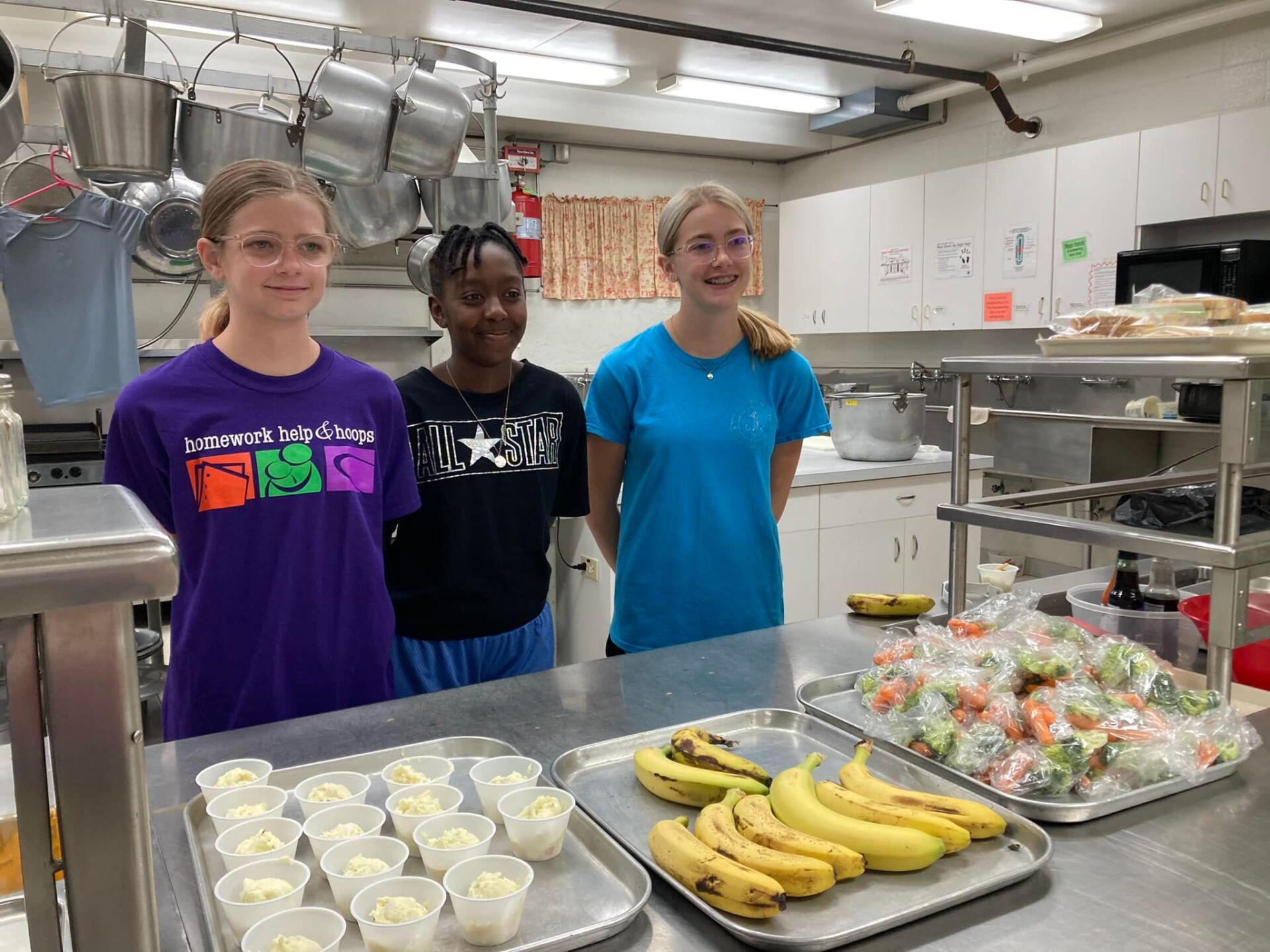 Three students stand in a kitchen behind trays of bananas, salad, and prepared food.