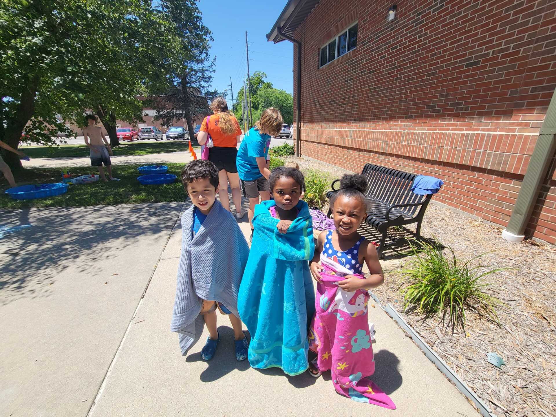 Three children in colorful clothes walking on a sunny sidewalk beside a brick building.