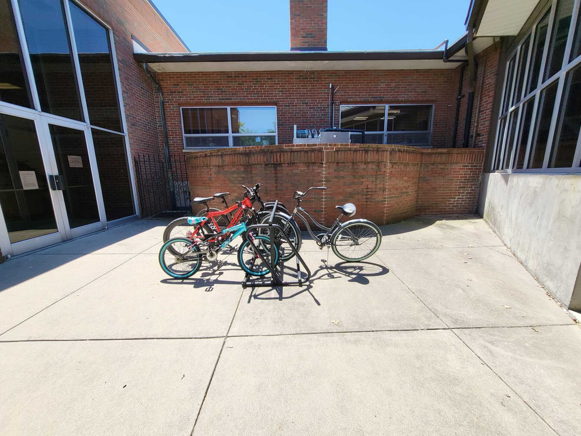 Bicycles parked in a sunny school courtyard beside brick buildings.
