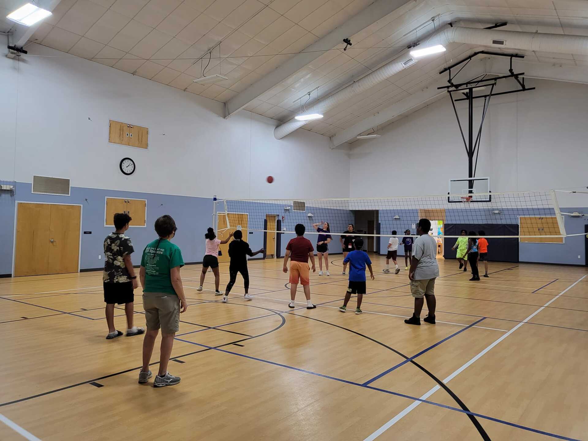 People playing indoor volleyball in a gym, with a net across the court and players spread out.