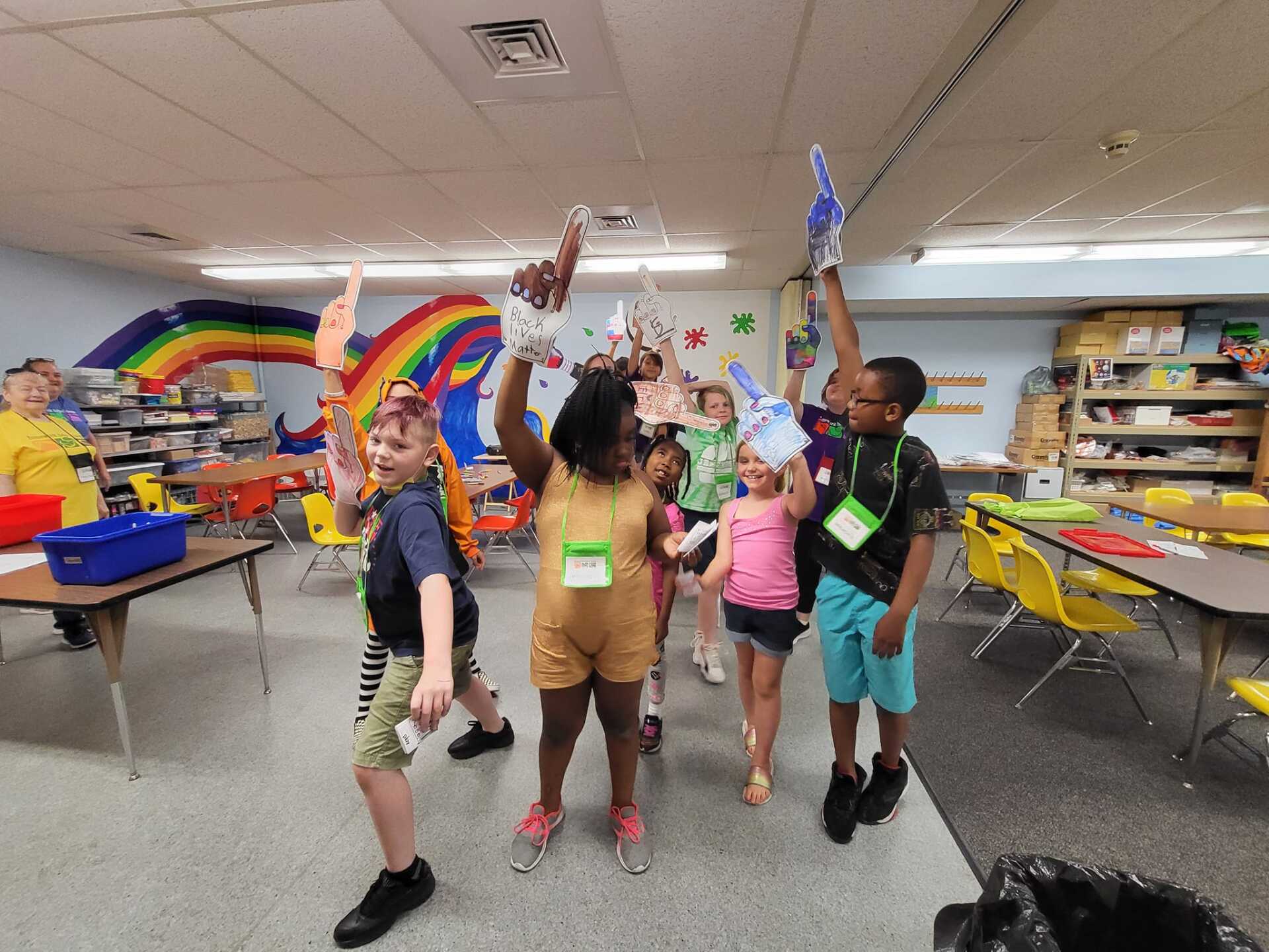 Children celebrating in a classroom, raising their hands amid rainbow decorations and tables.