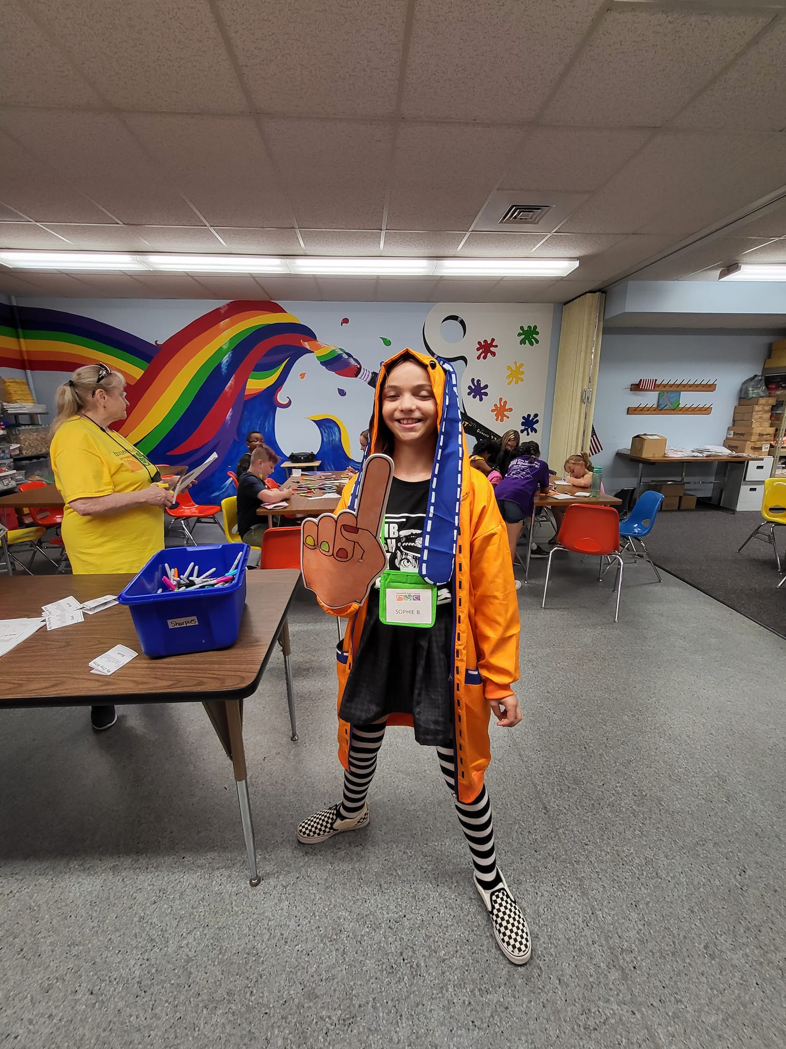 Person in a Halloween costume stands in a classroom decorated with a rainbow mural and spooky decorations.