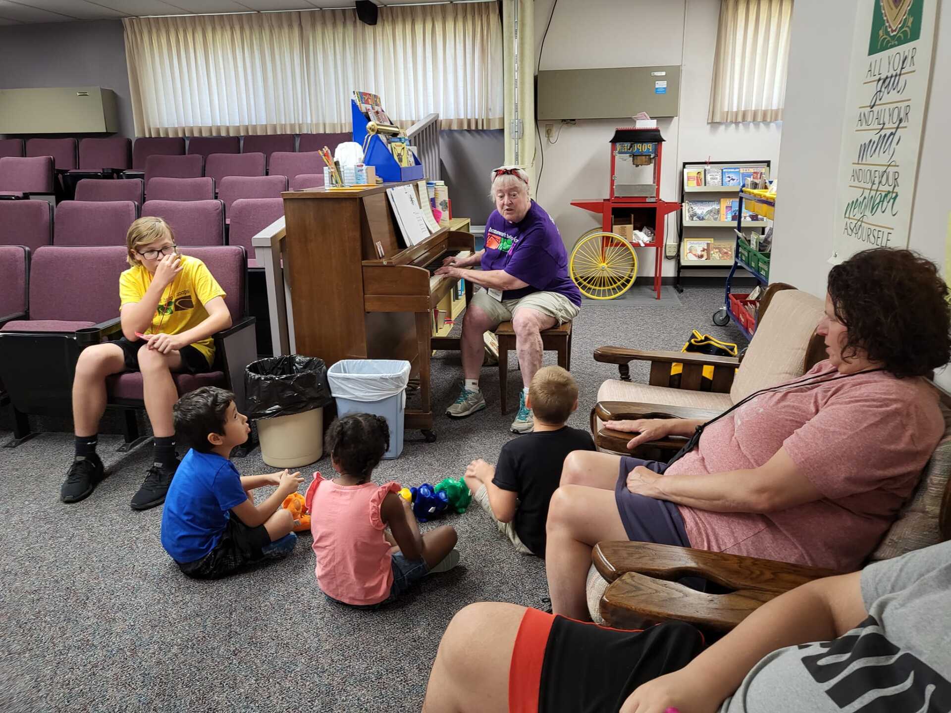 Small group sitting on carpet in a room, with a person at an upright piano leading a gathering.