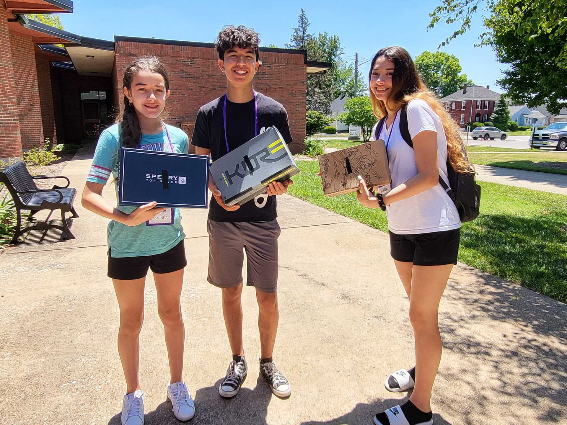 Three students standing outdoors, smiling and holding laptops on a sunny campus walkway.