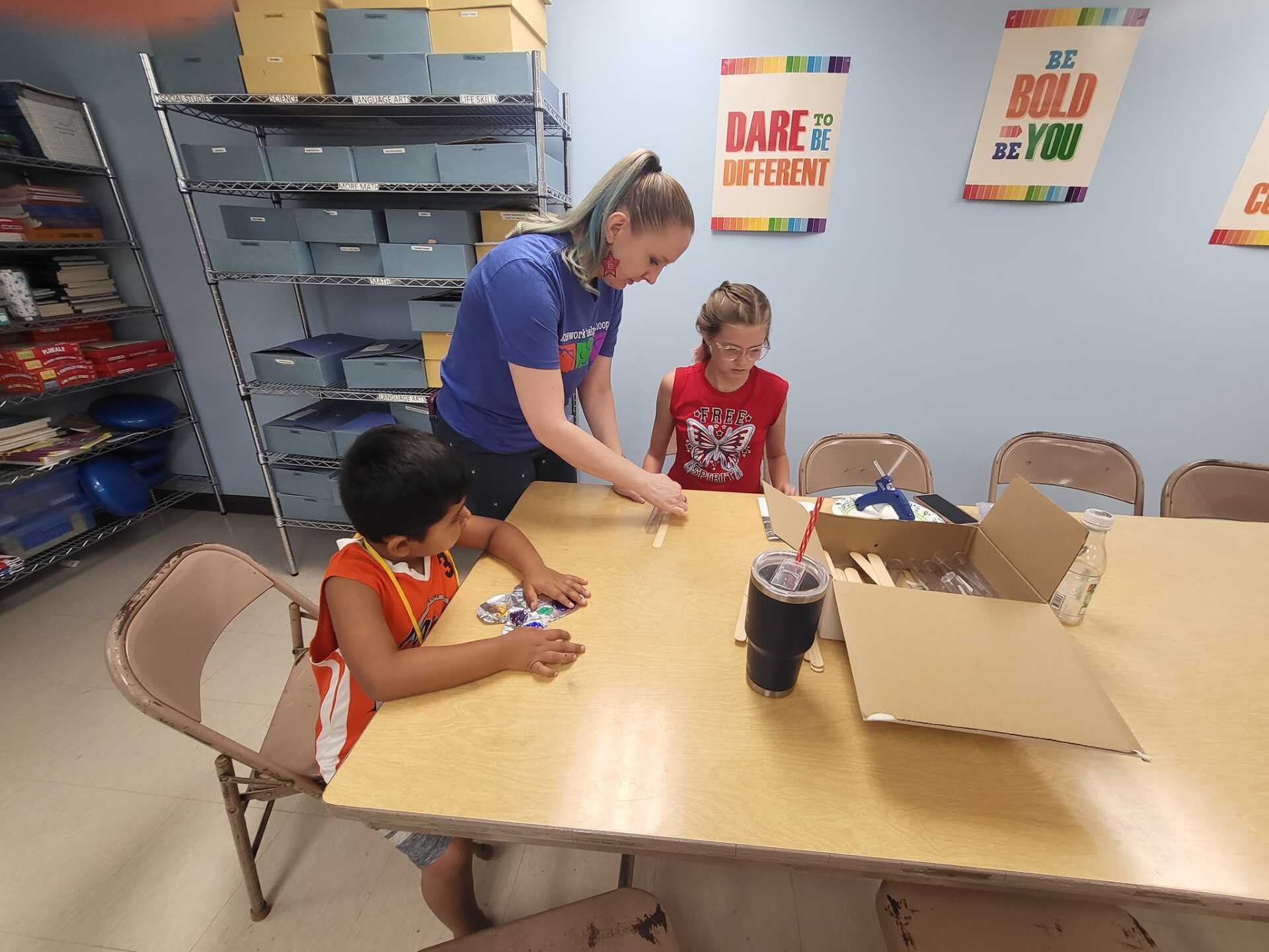 Adult helping two children with a craft project at a table in a classroom-like room