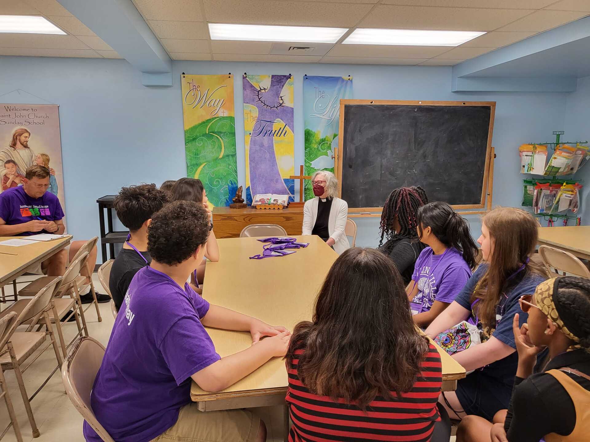 Students in a classroom sit in a circle facing a teacher near a chalkboard and colorful wall art.