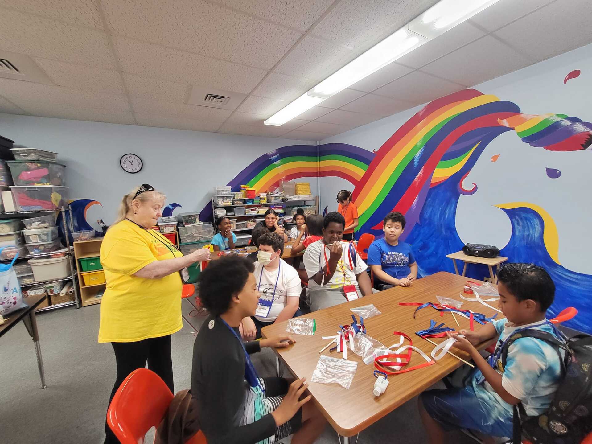 Teacher speaks to seated students in a classroom with colorful rainbow murals and craft supplies on tables