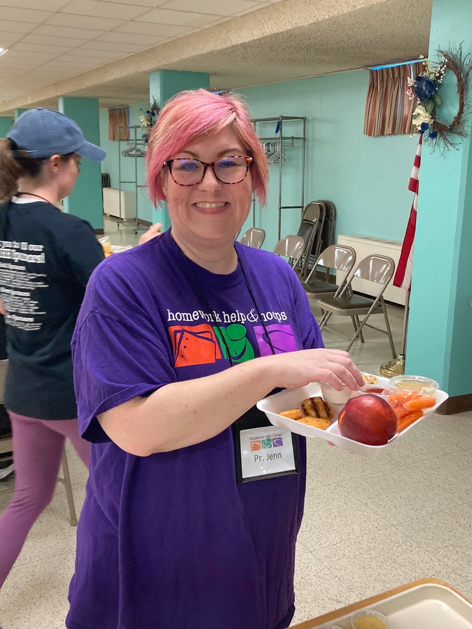 Person with pink hair and glasses holding a tray of food in a cafeteria-like room
