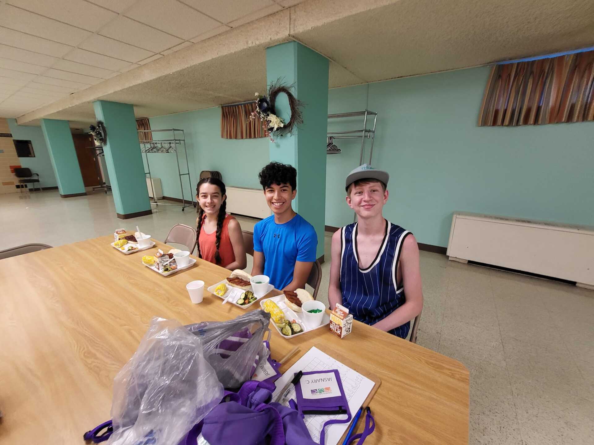 Three people seated at a long table in a bright hall, with food and drinks in front of them.