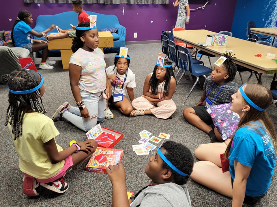Children sit in a circle on a classroom floor, playing a card game with an adult nearby.