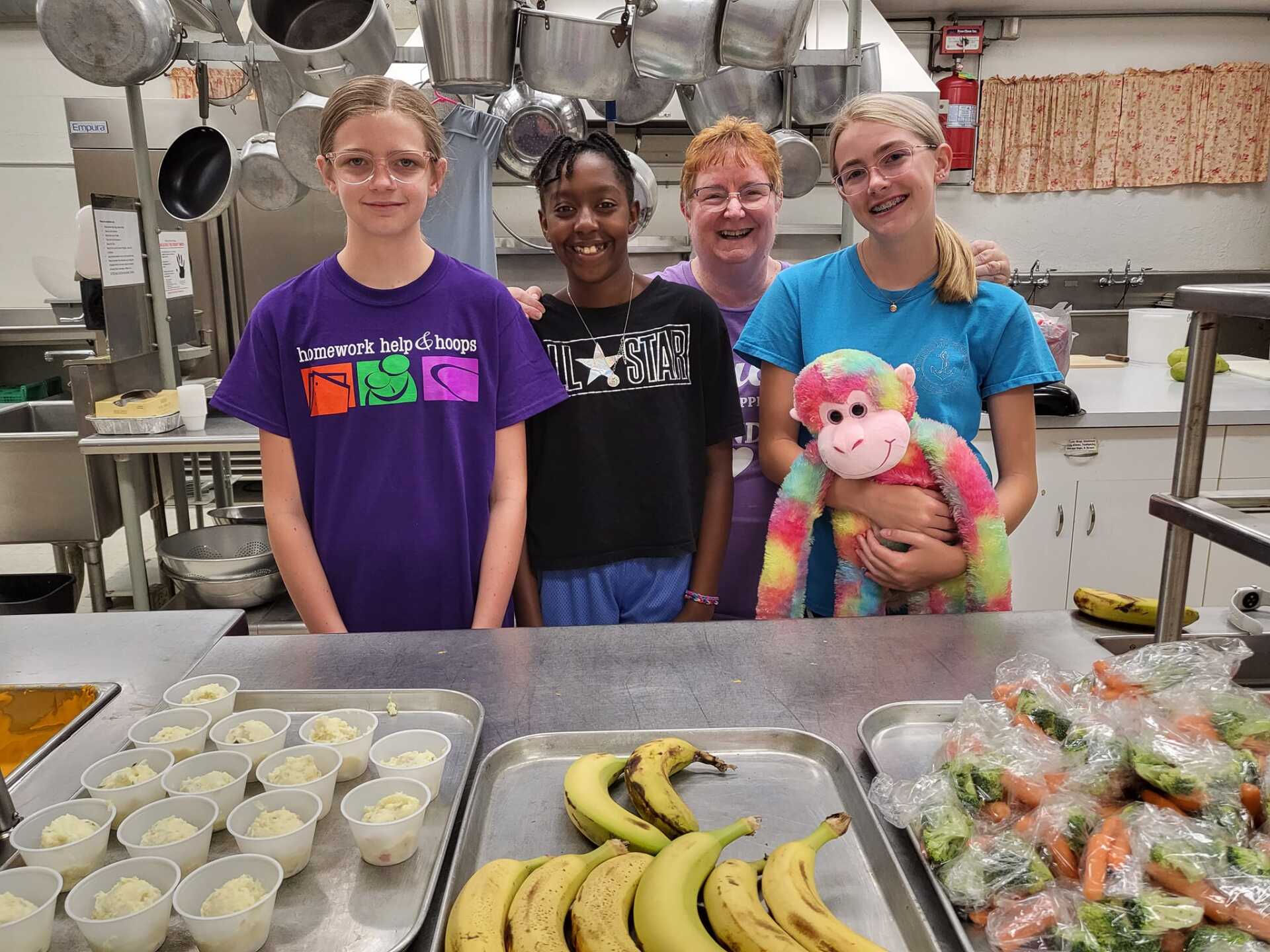 Four people standing behind trays of food in a kitchen, smiling and holding a stuffed toy.