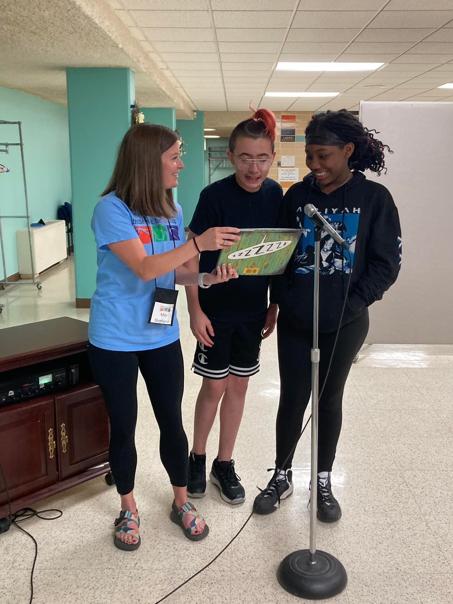 Three people stand in a hallway, smiling and reading a green box near a microphone stand.