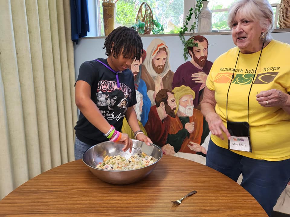 Two people mixing food in a large bowl at a wooden table, with a mural in the background.