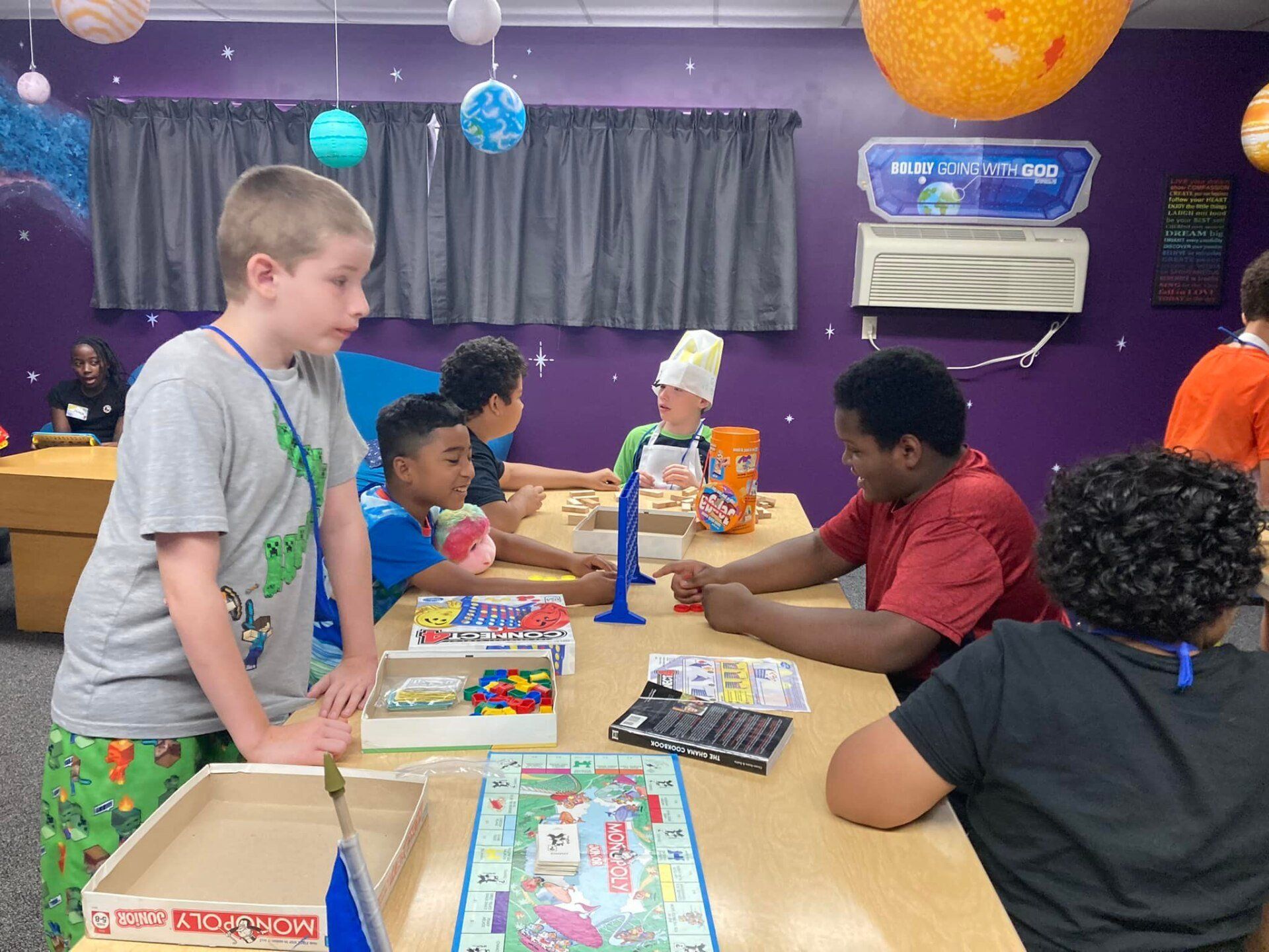 Children play a board game at a table in a purple game room with balloons and an air conditioner.
