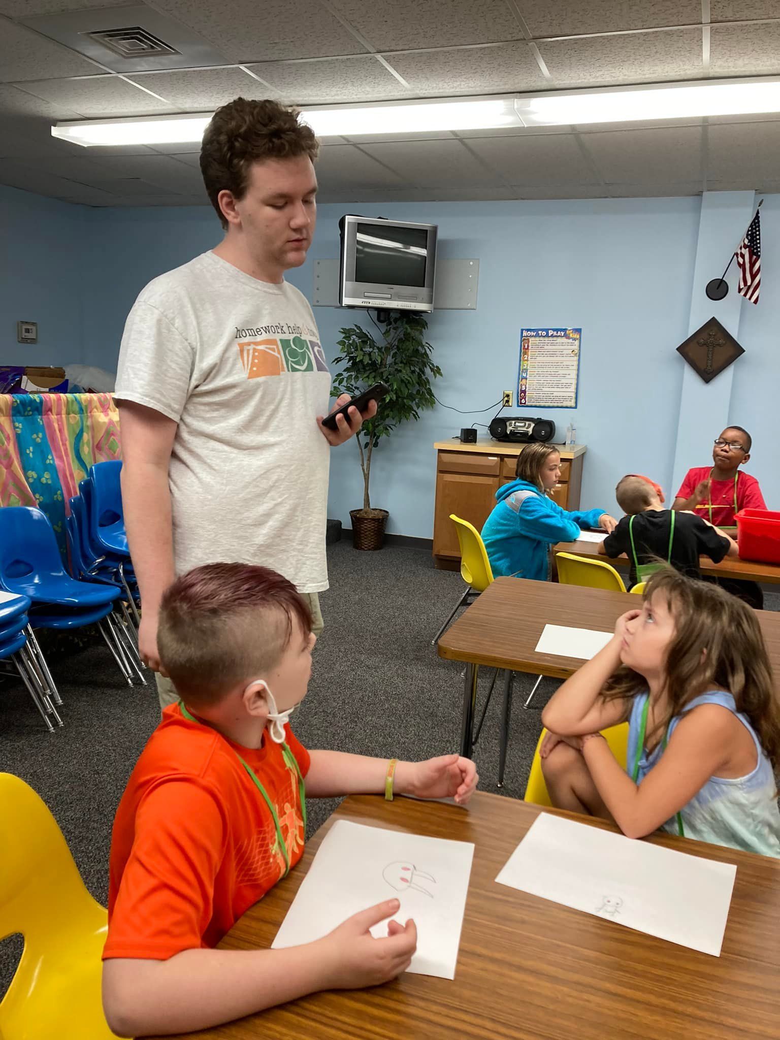 Man standing beside seated children in a classroom, with tables and papers in the foreground.