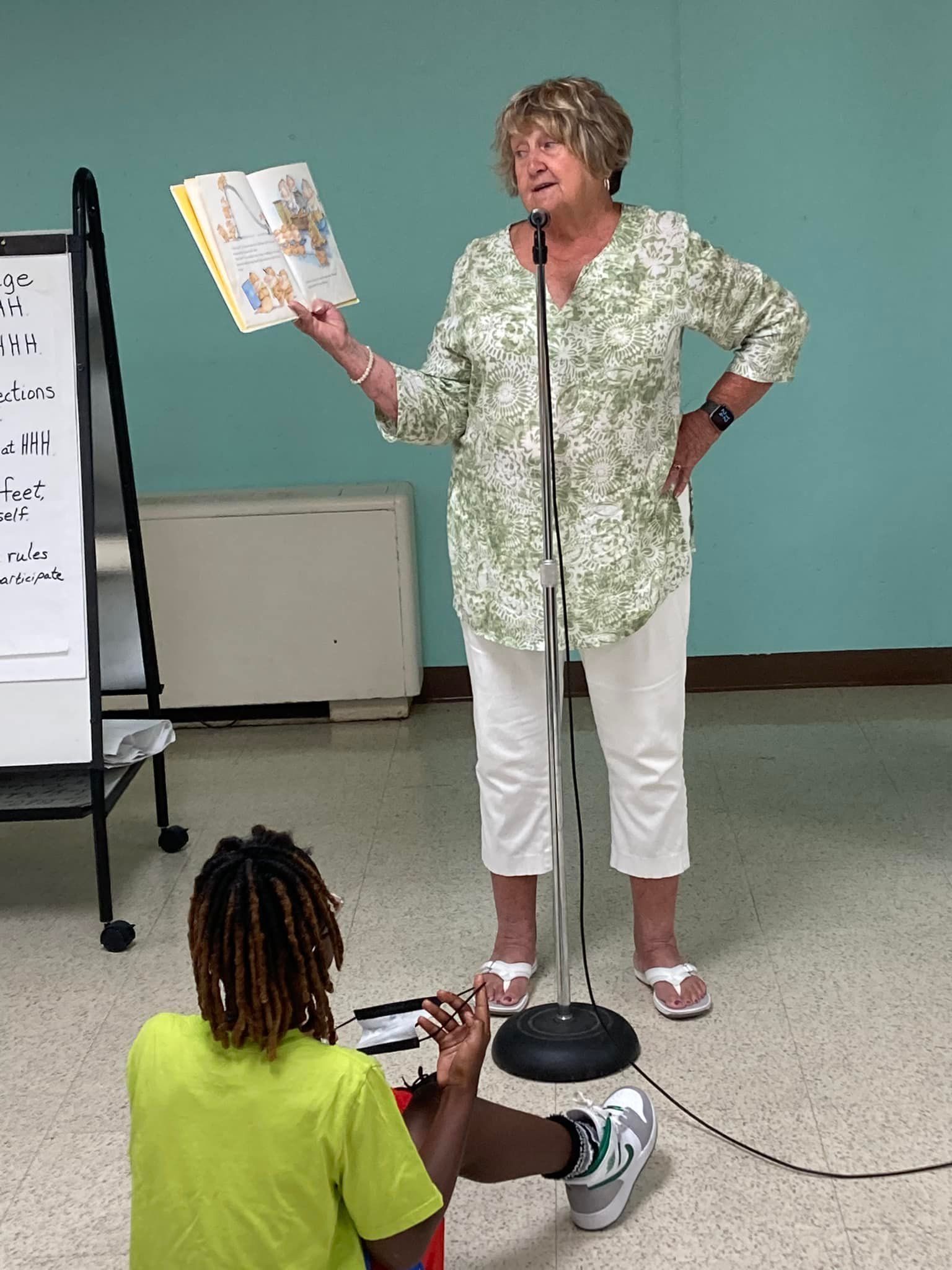 Woman speaking at a microphone while holding an open book, with a seated child in the foreground.