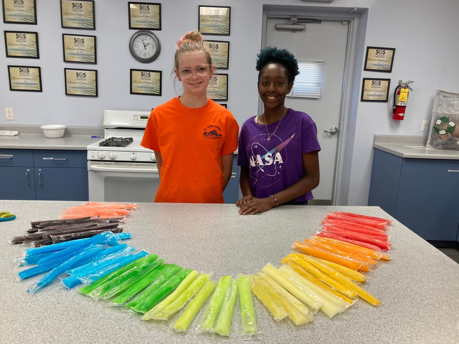Two smiling people stand behind a table with rainbow-colored plastic items arranged in a fan.