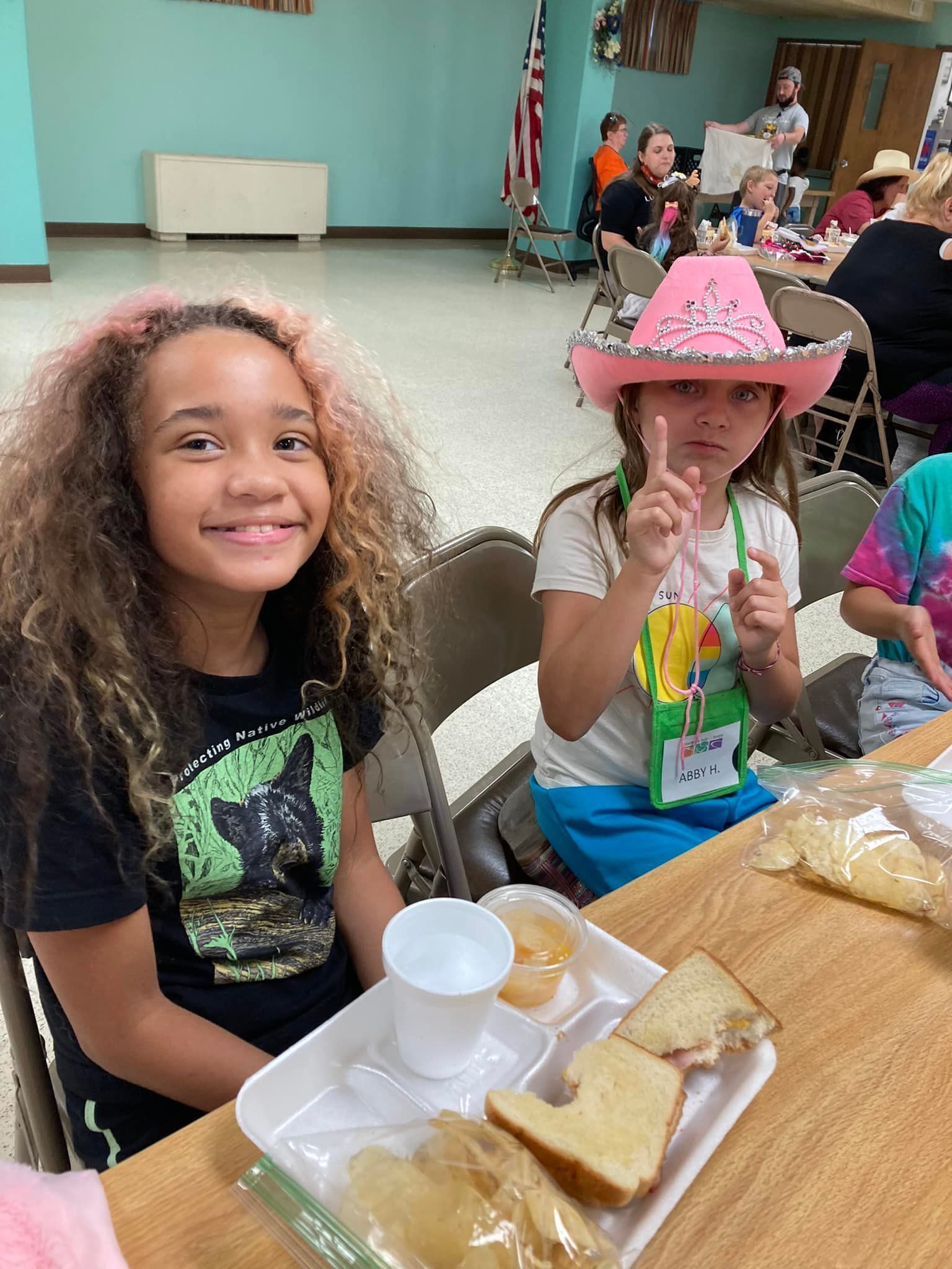 Two children smiling at a table with plates and cups in a cafeteria, one wearing a pink hat