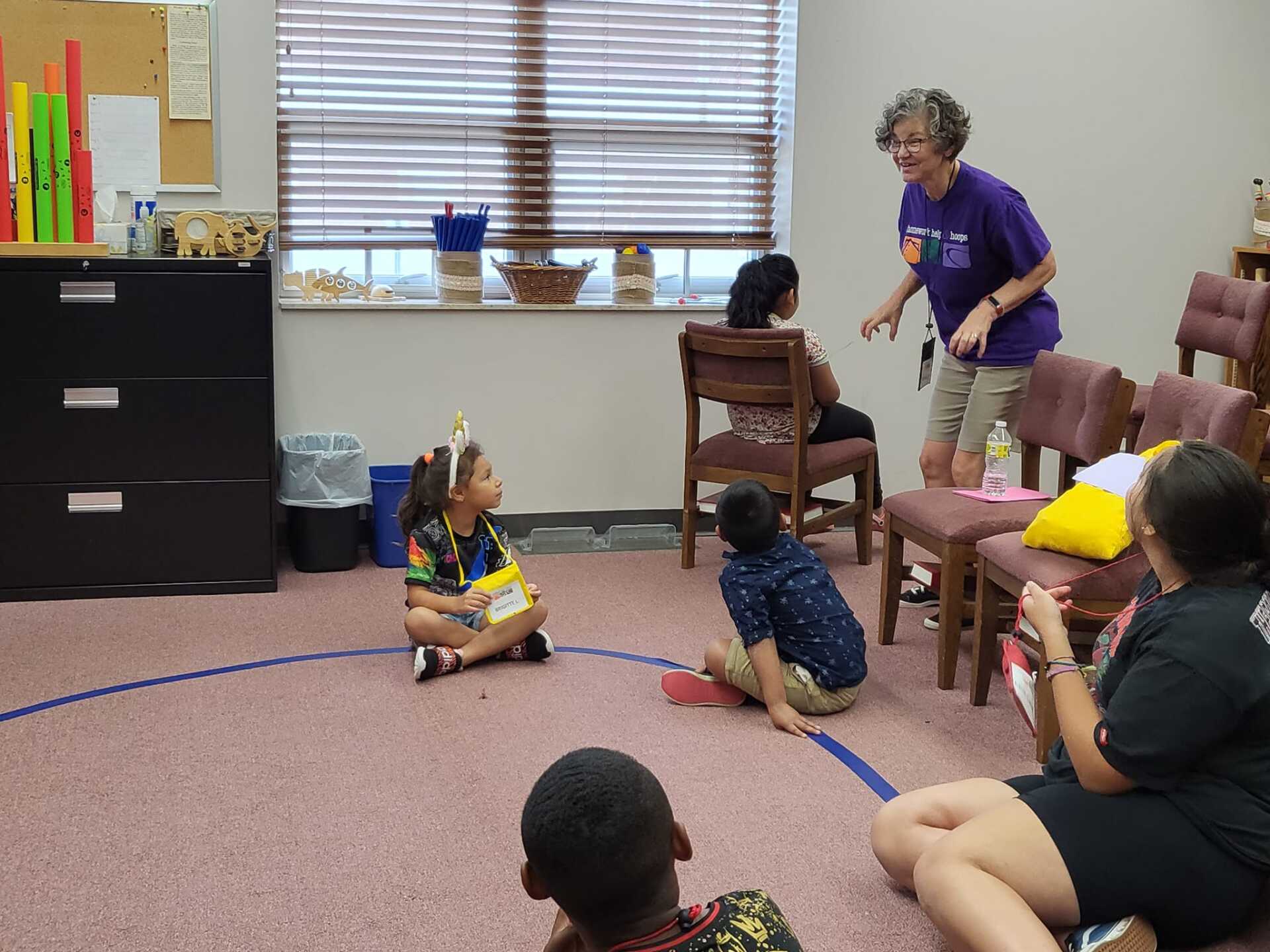 Children in a classroom circle while a woman stands nearby, with toys and chairs around them.