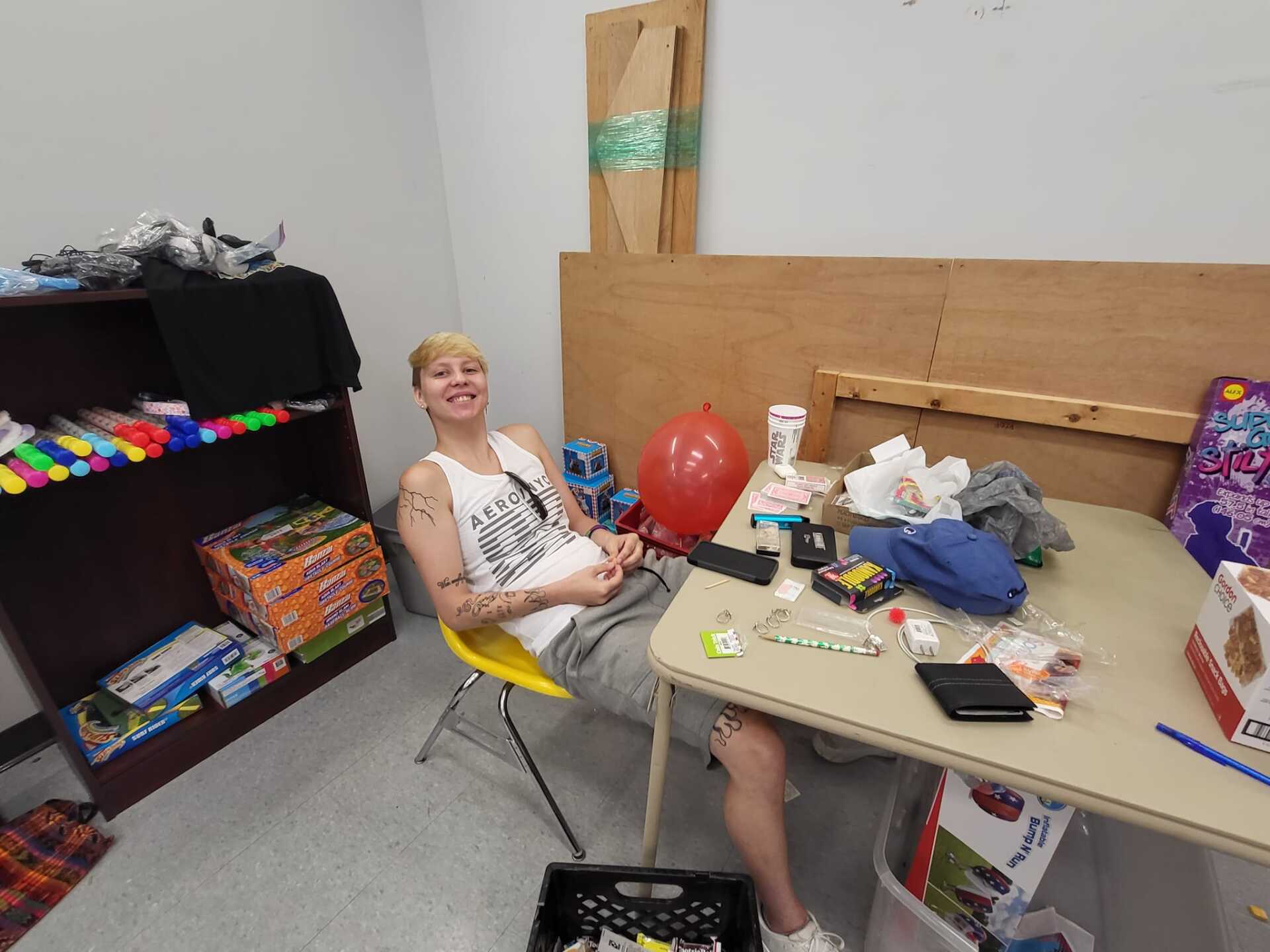 Person smiling at a cluttered table in a room with toys, a red balloon, and colorful supplies.