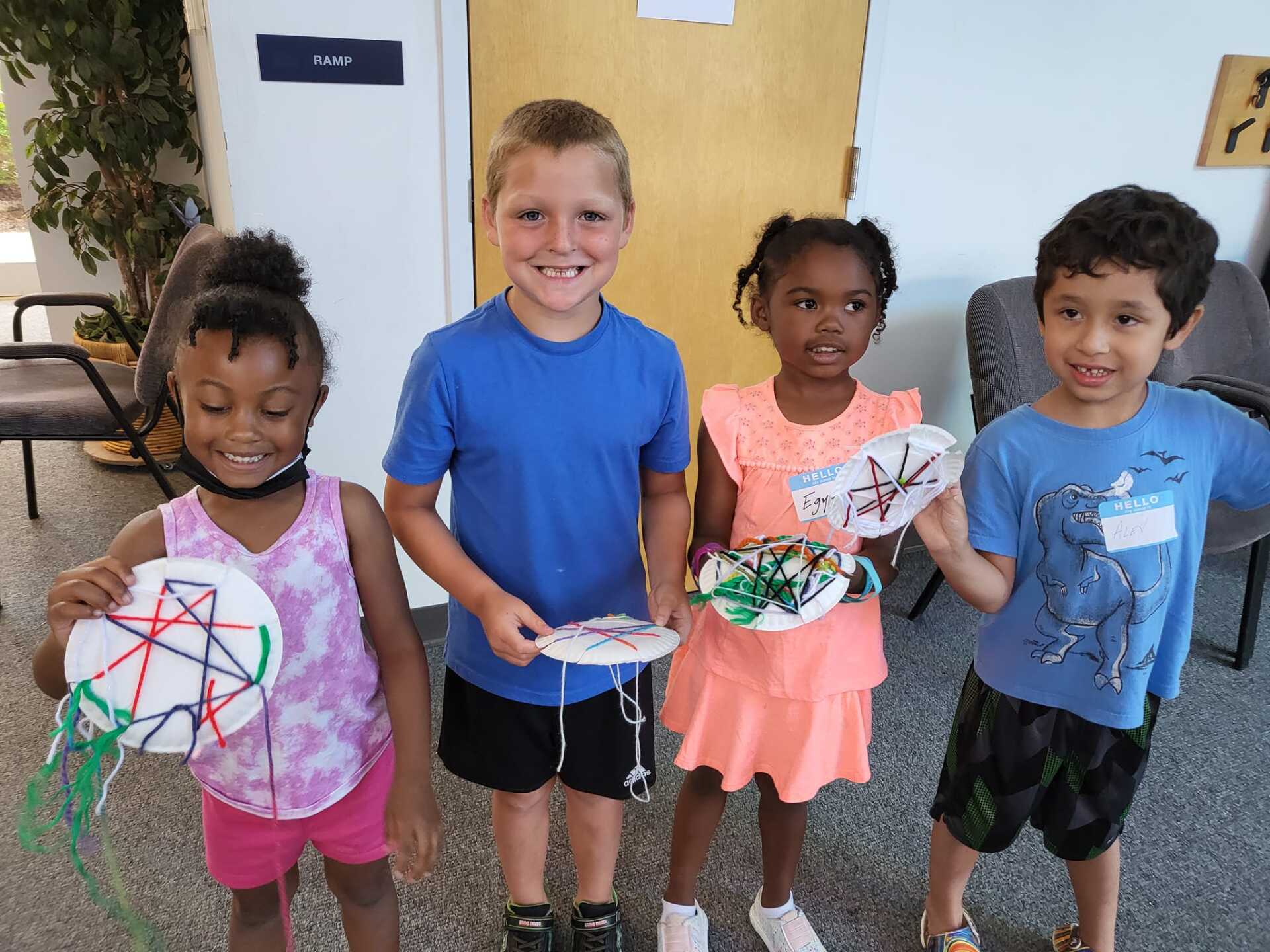 Four smiling children hold paper crafts while standing indoors against a white wall.