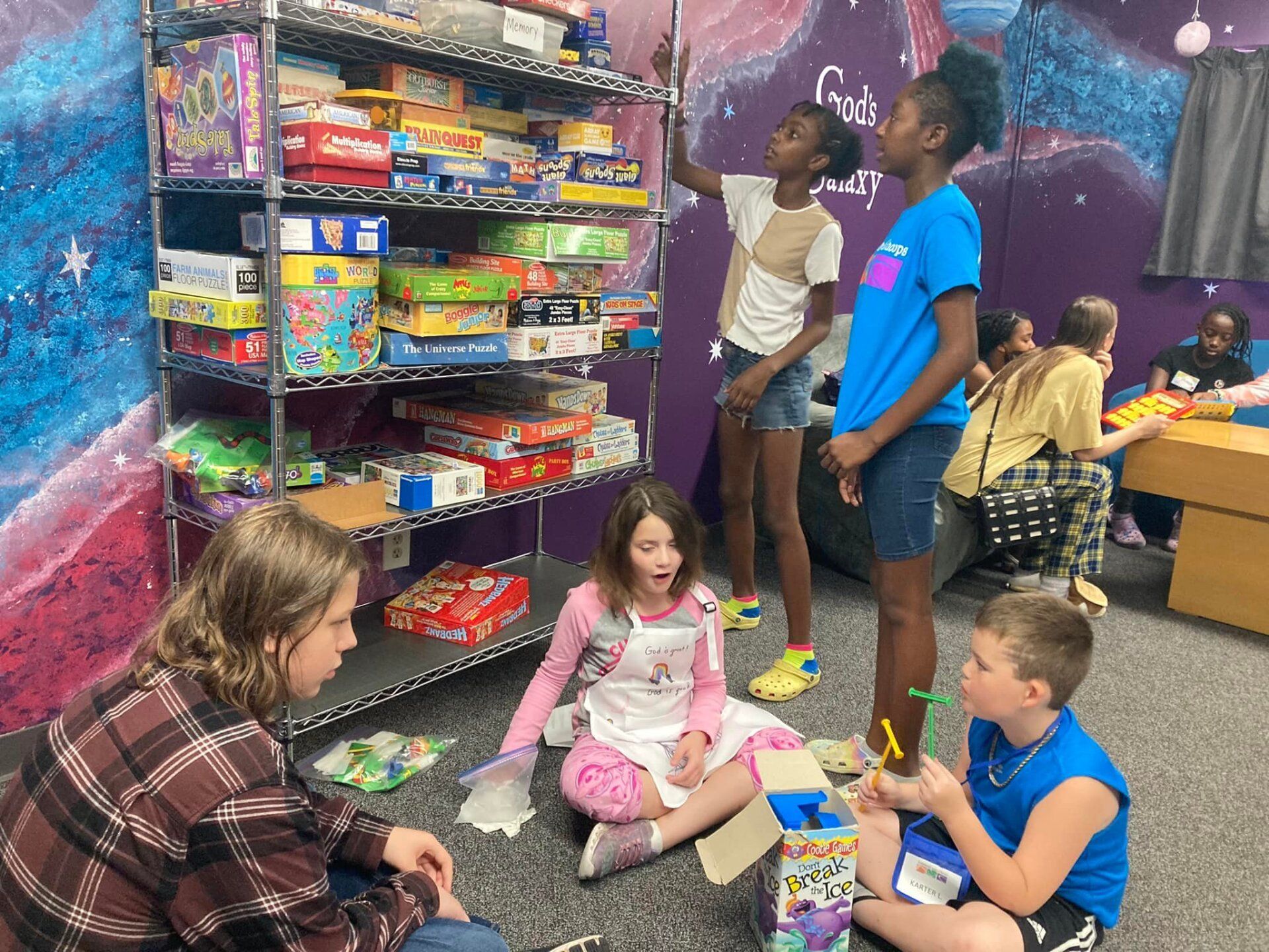 People sitting on the floor in a colorful playroom, surrounded by shelves of toys and books.