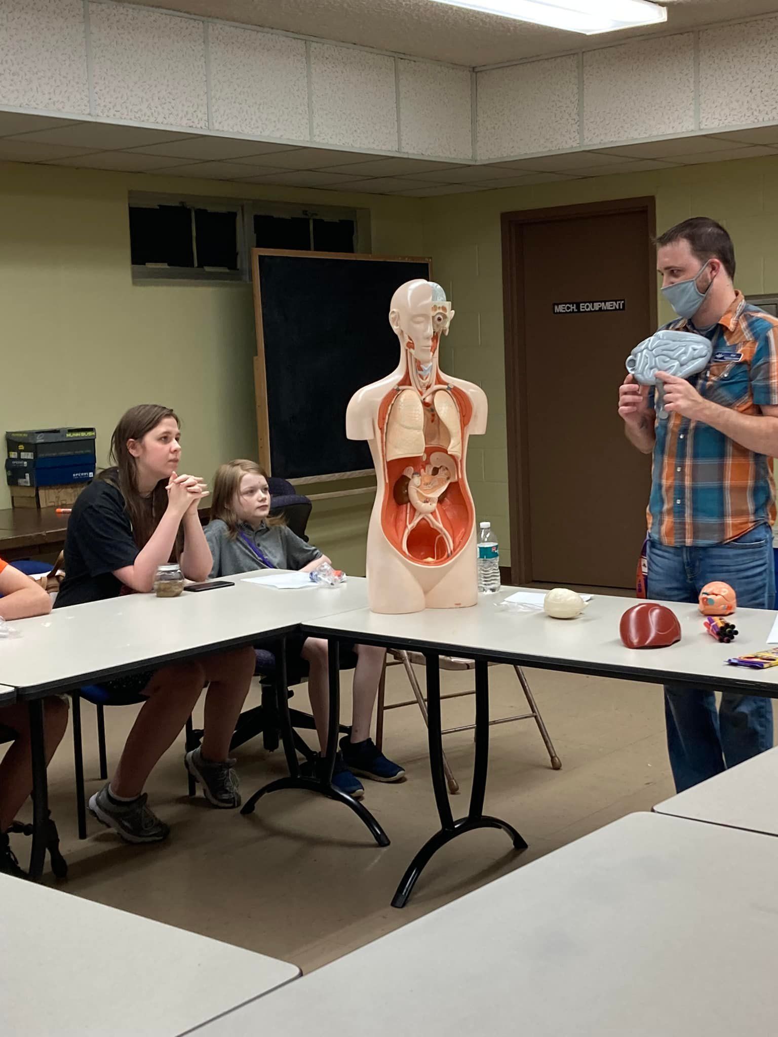 Classroom anatomy demo with torso model, seated students, and a man holding a baby doll.